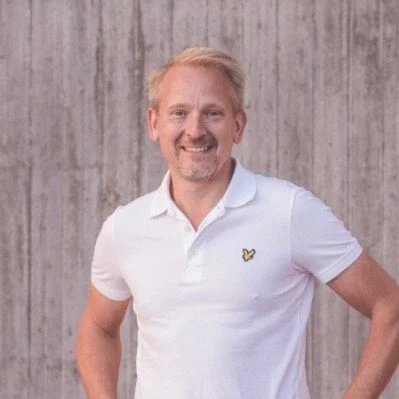 A smiling man with light hair and a beard wearing a white polo shirt standing in front of a wooden background.