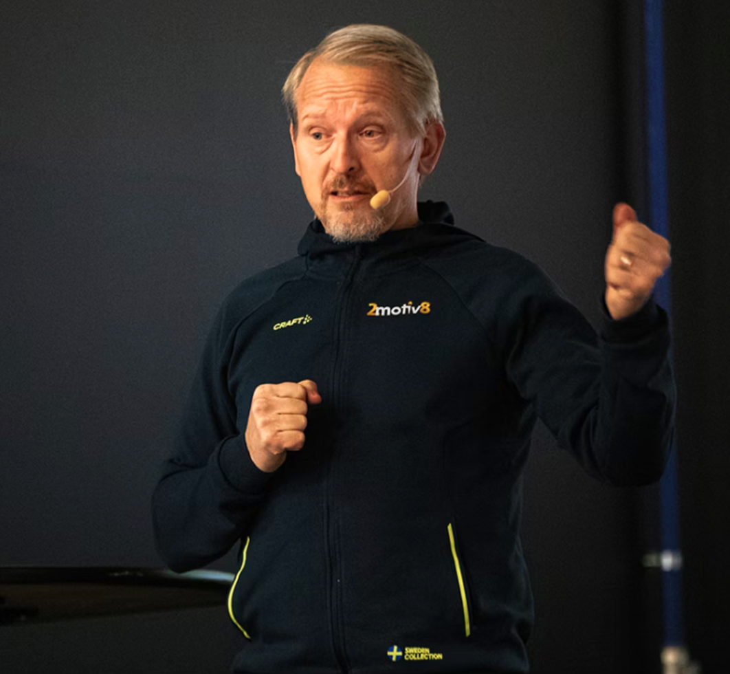 A middle-aged man with light hair and beard wearing a black athletic jacket with yellow accents, speaking into a headset microphone, and gesturing with his right hand during a presentation in a dark room.