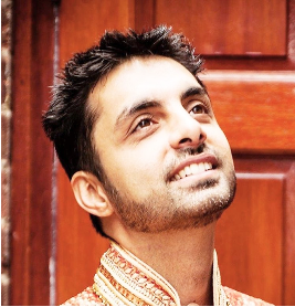 A man with short dark hair and facial hair is smiling and looking upward, standing in front of a wooden background.