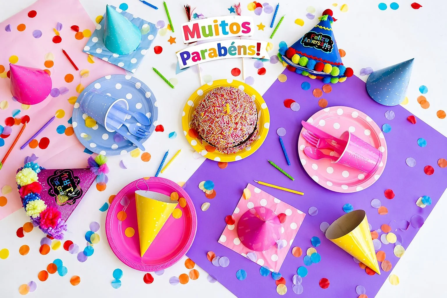 Decoração de festa de aniversário infantil com bolo colorido, chapéus de festa, confettis e mensagem “Muitos Parabéns”, ambiente alegre e festivo para crianças