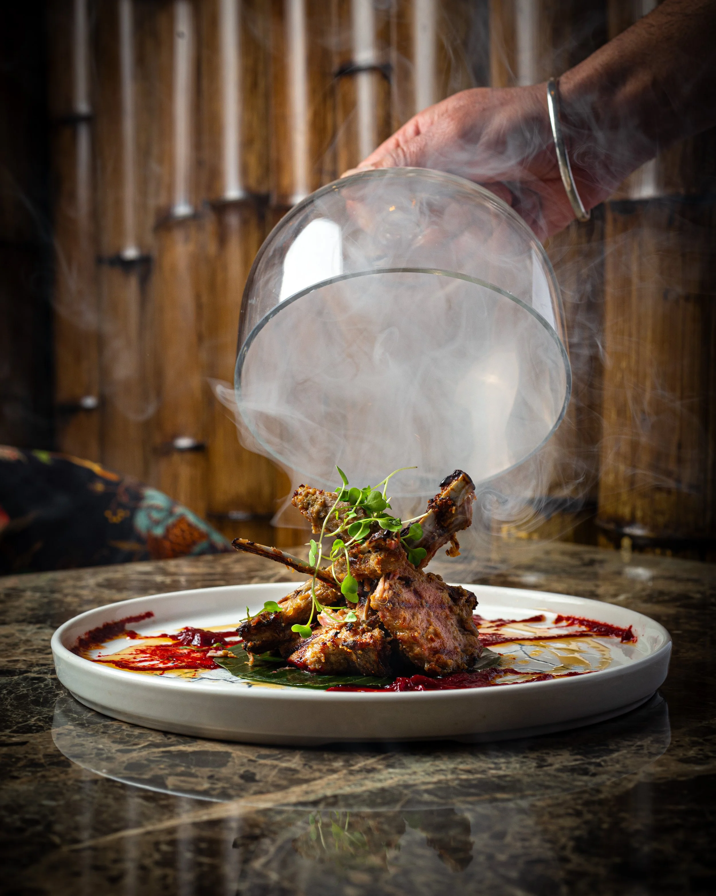 Smoke cloche being lifted from charcoal-grilled tandoori lamb chops on a marble plate