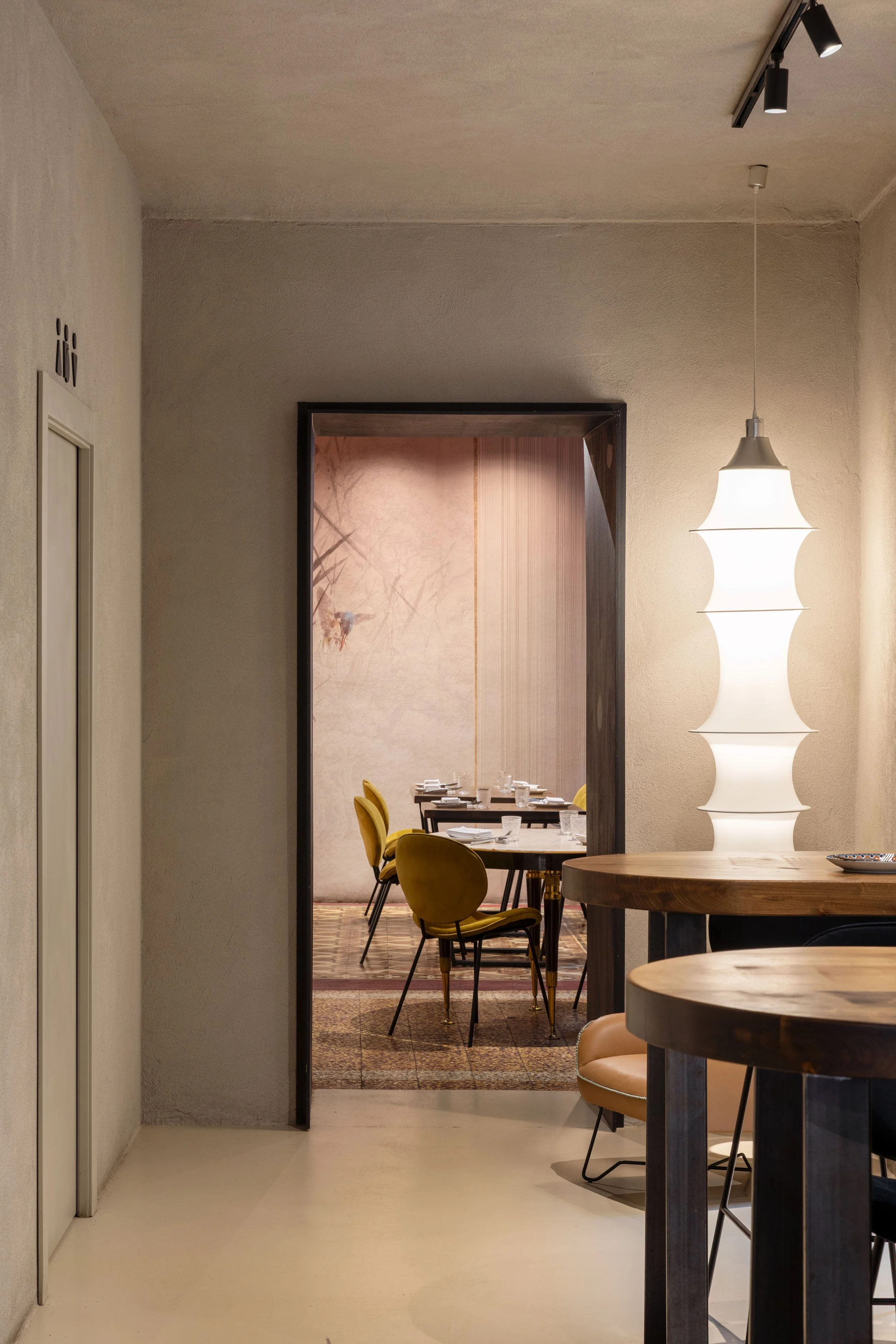 corridoio di collegamento tra le sale principali del ristorante, con tavoli alti su misura e luce scultorea che da personalità a questo spazio neutro. I portali in legno verniciato nero segnano l'accesso alle sale.