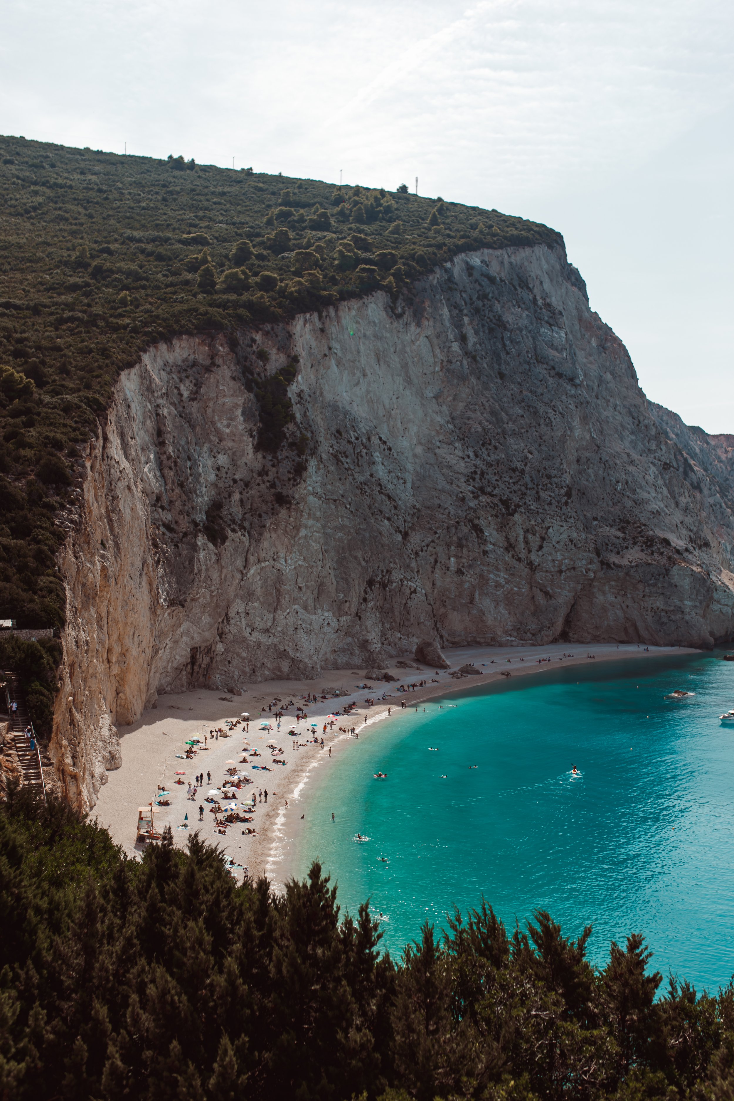 A beach with turquoise water, a sandy shore, and people sunbathing and swimming, adjacent to tall, rocky cliffs and lush green vegetation.