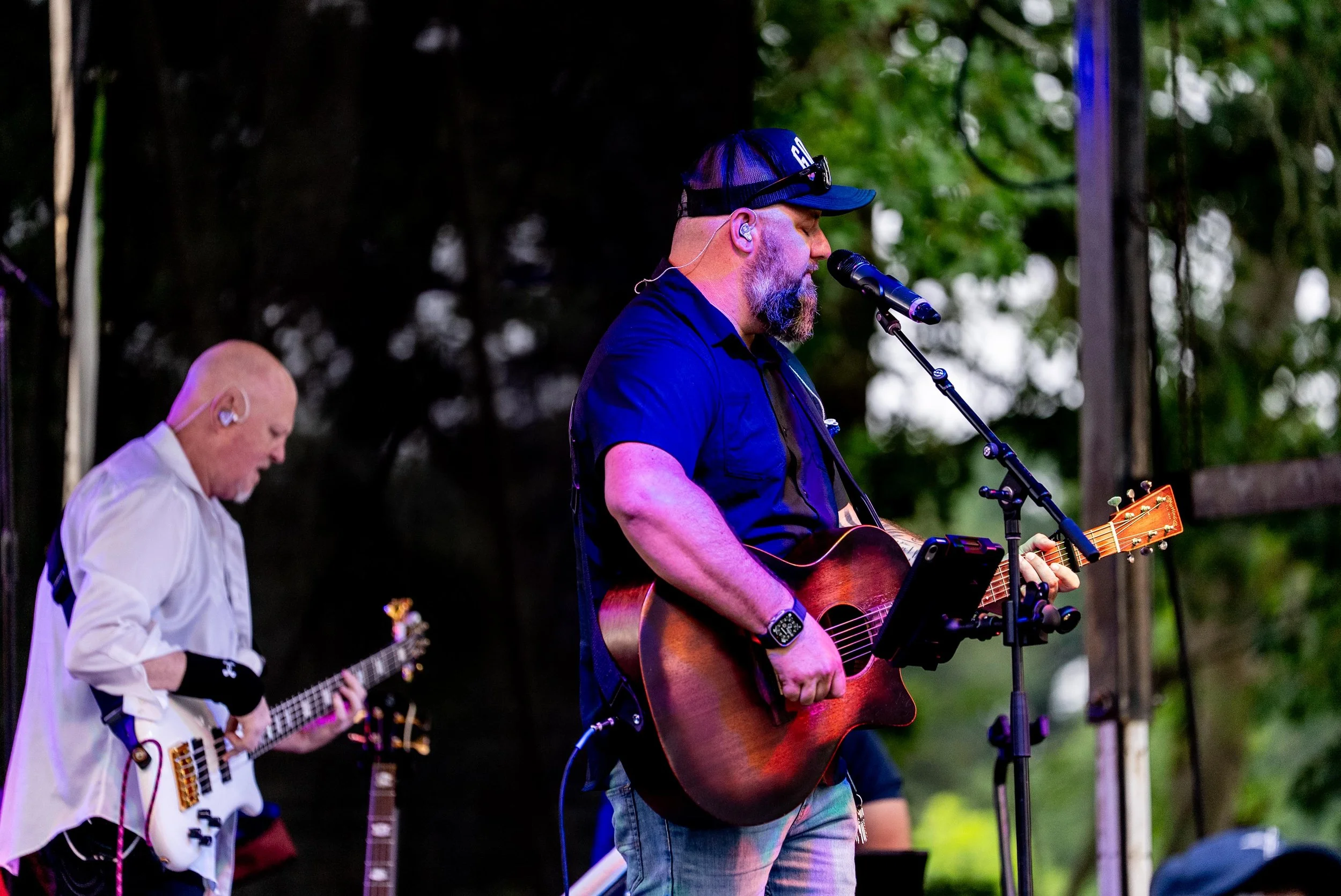 Two male musicians performing on stage outdoors. The one in the foreground is playing an acoustic guitar and singing into a microphone. He has a beard, is wearing a black cap, and a black shirt. The musician in the background is playing an electric g