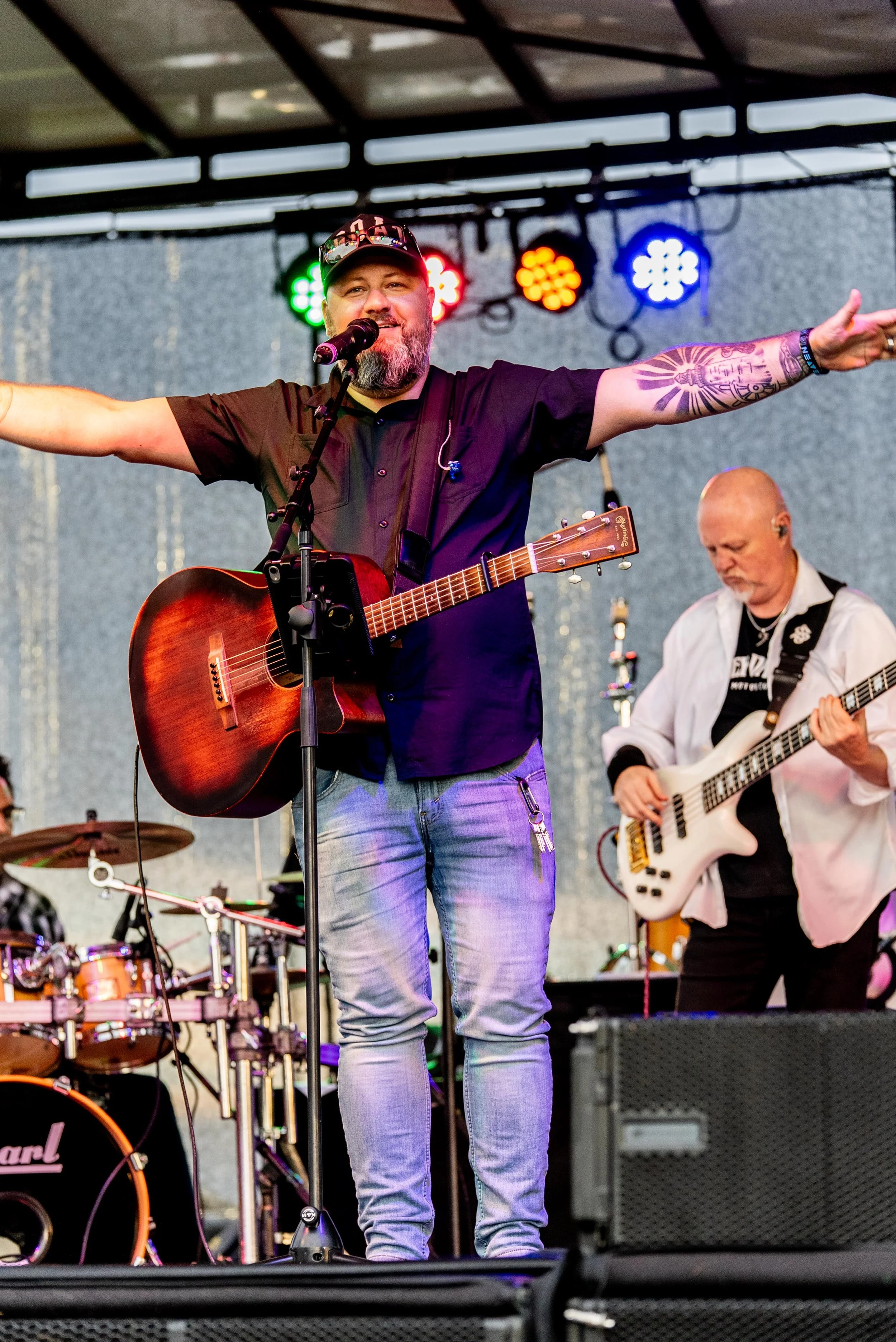A man with a beard and a cap plays an acoustic guitar and sings into a microphone on stage, with arms outstretched, during a live music performance. A drummer and another guitarist are visible in the background, with stage lights overhead.