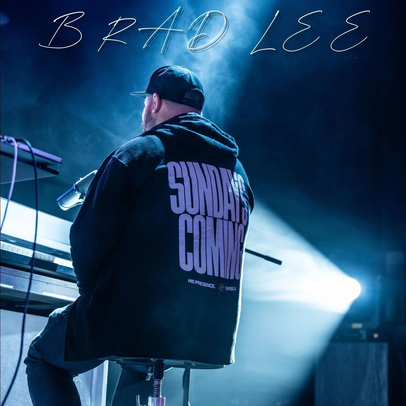 A man with a beard wearing a black hoodie with purple text that says 'SUNDAYS COMING' and a black cap, sitting at a keyboard on stage backstage with blue lighting and smoke effects in the background.