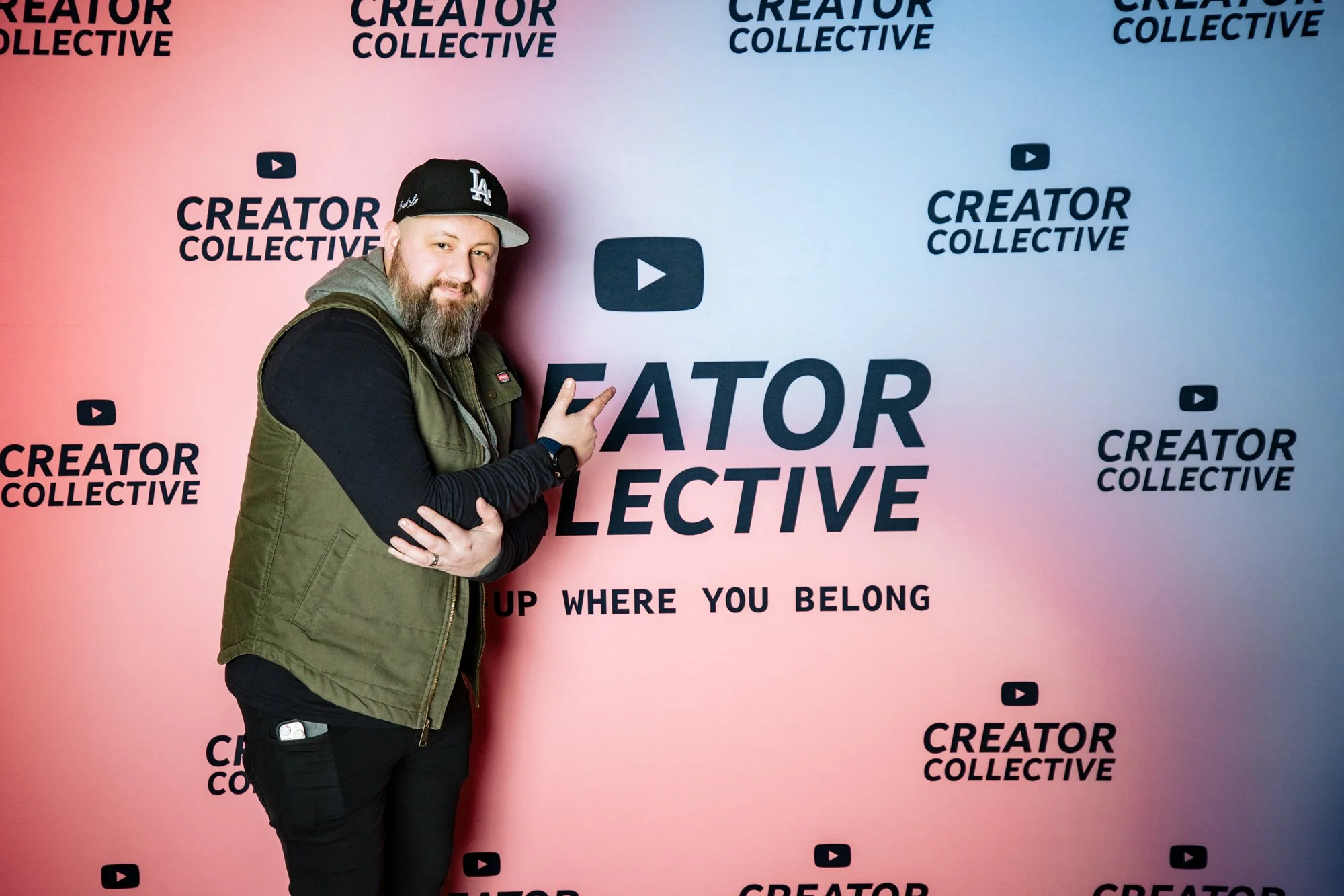 A man with a beard wearing a black baseball cap, black long sleeve shirt, and an olive green vest, standing in front of a backdrop with the words "Creator Collective" and the YouTube logo. He is pointing at the text on the backdrop and smiling.