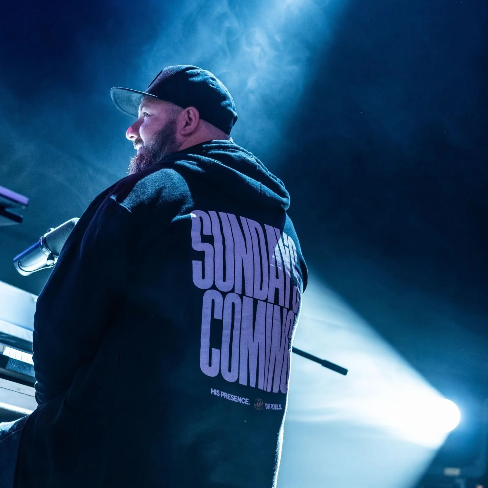 A man with a beard wearing a black hoodie and a black cap, sitting at a piano on stage, with a blue spotlight and haze around him, seen from the back and side.