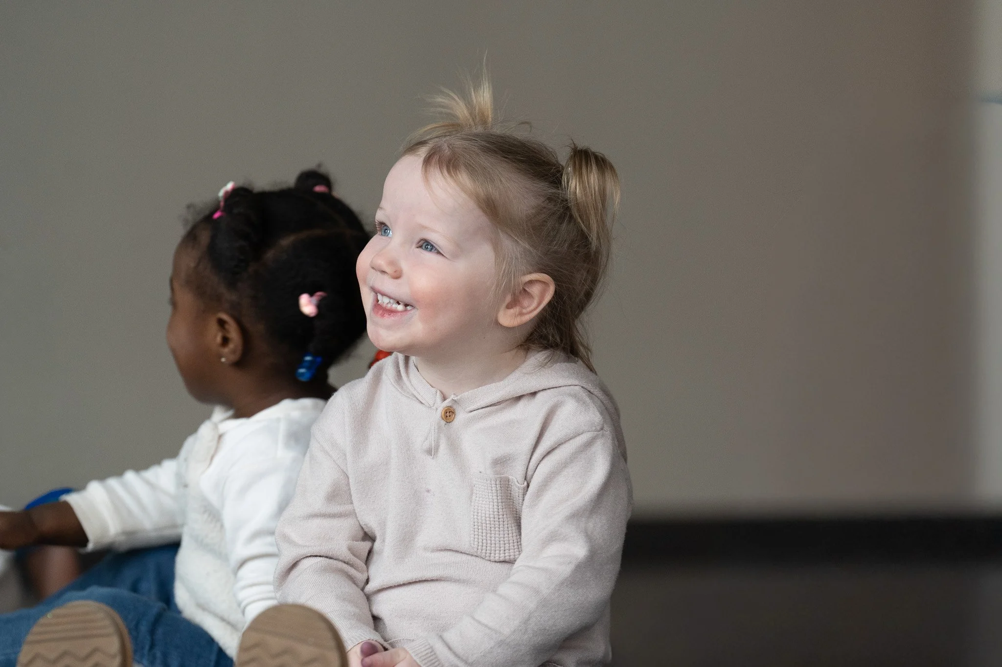 A smiling young girl with blonde hair tied in pigtails sitting on the floor next to a girl with dark hair styled in braids. Both are indoors.