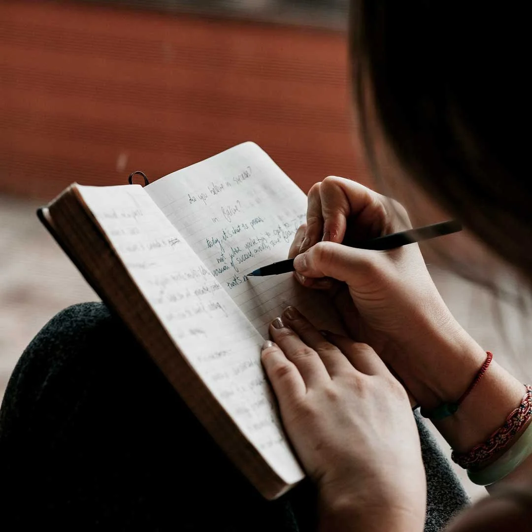 A person writing in a journal, viewed from over the shoulder.