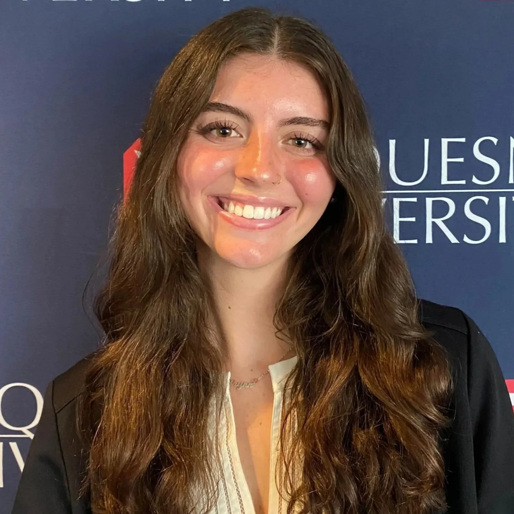 A young woman with long wavy brown hair, light skin, gray eyes, a nose piercing, and wearing a black jacket over a cream-colored shirt, smiling in front of a blue background with white text.