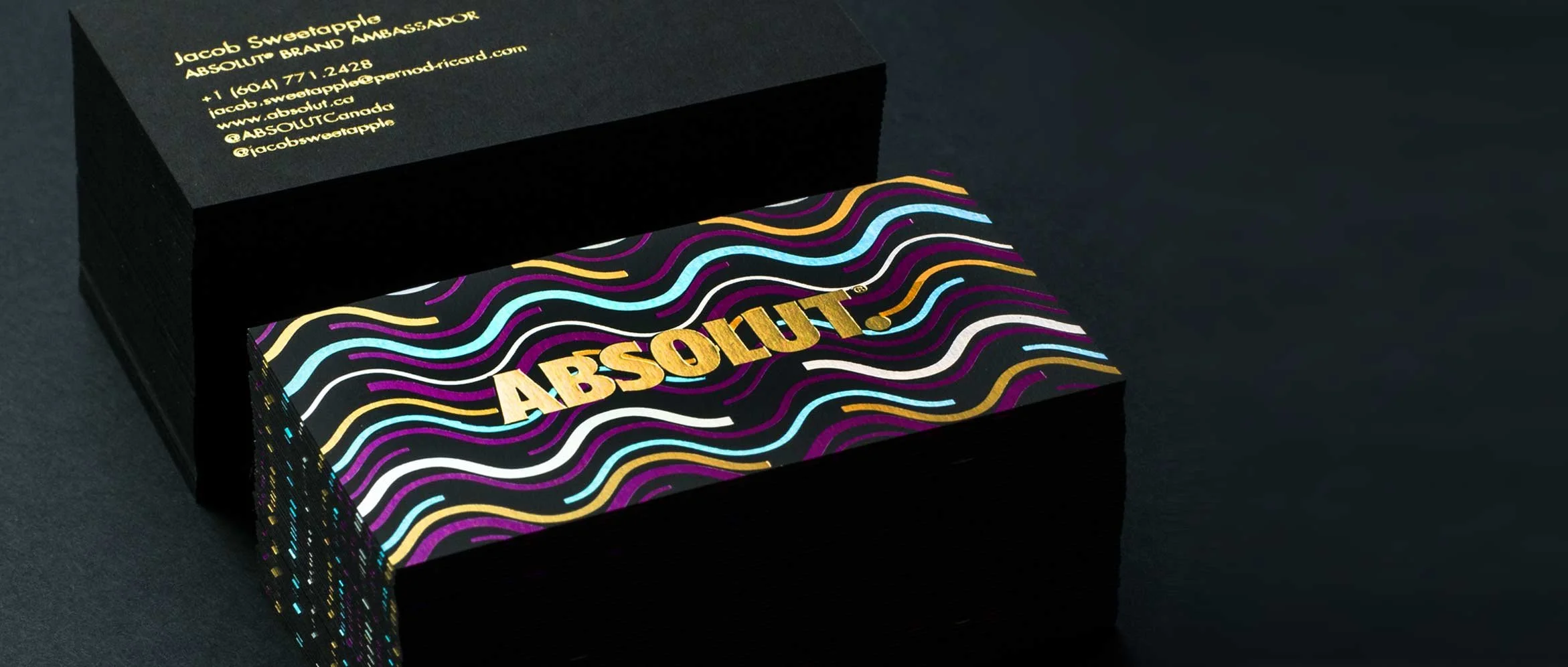 A black box with gold print displaying contact information for Jacob Sweatapple, an ambassador for ABSOLUT. The box is partially open, revealing a vibrant, colorful set of business cards with a wavy pattern and the word "ABSOLUT" in gold letters on a black background.