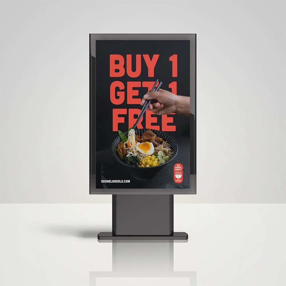 Digital advertisement display for a promotion by Doo Noodle, featuring a bowl of ramen with a soft boiled egg, vegetables, and meat, and a hand holding chopsticks above it, with a large red text sign saying 'Buy 1 Get 1 Free'.