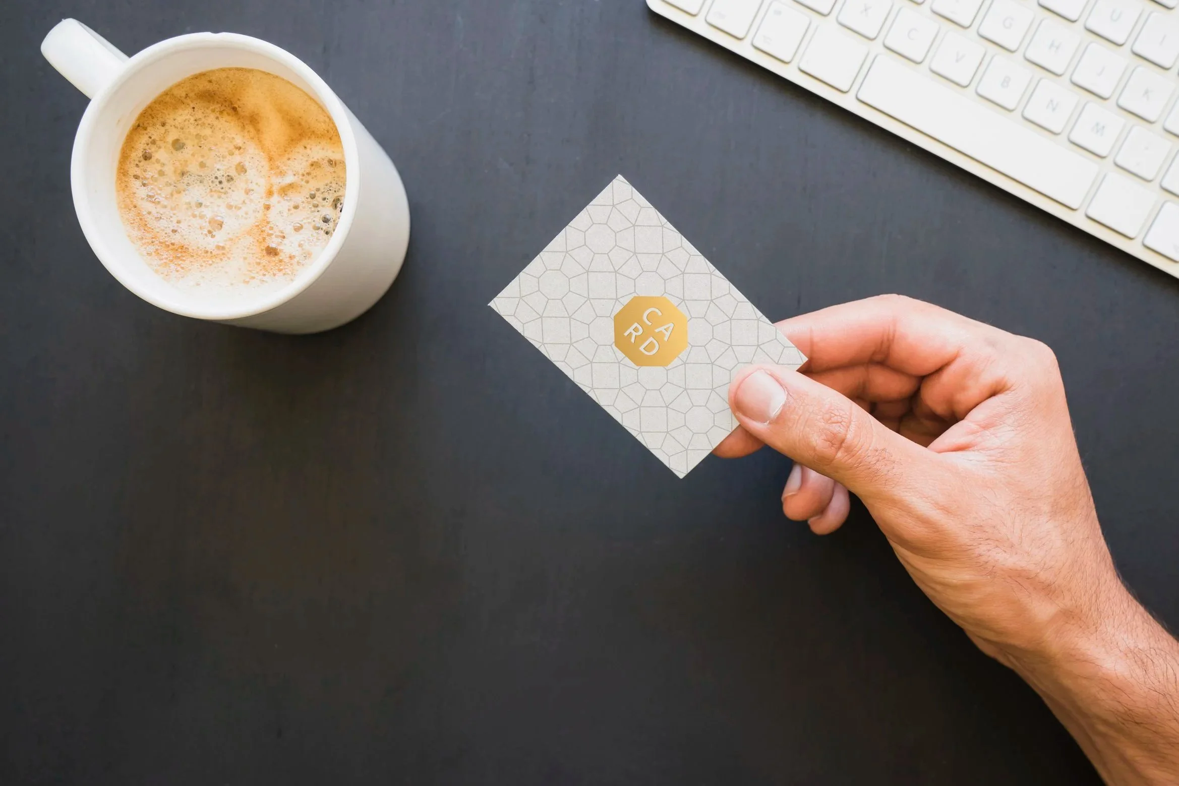 A hand holding a business card with a hexagonal logo that says 'CARD', a cup of coffee, a computer keyboard, and a black desk.
