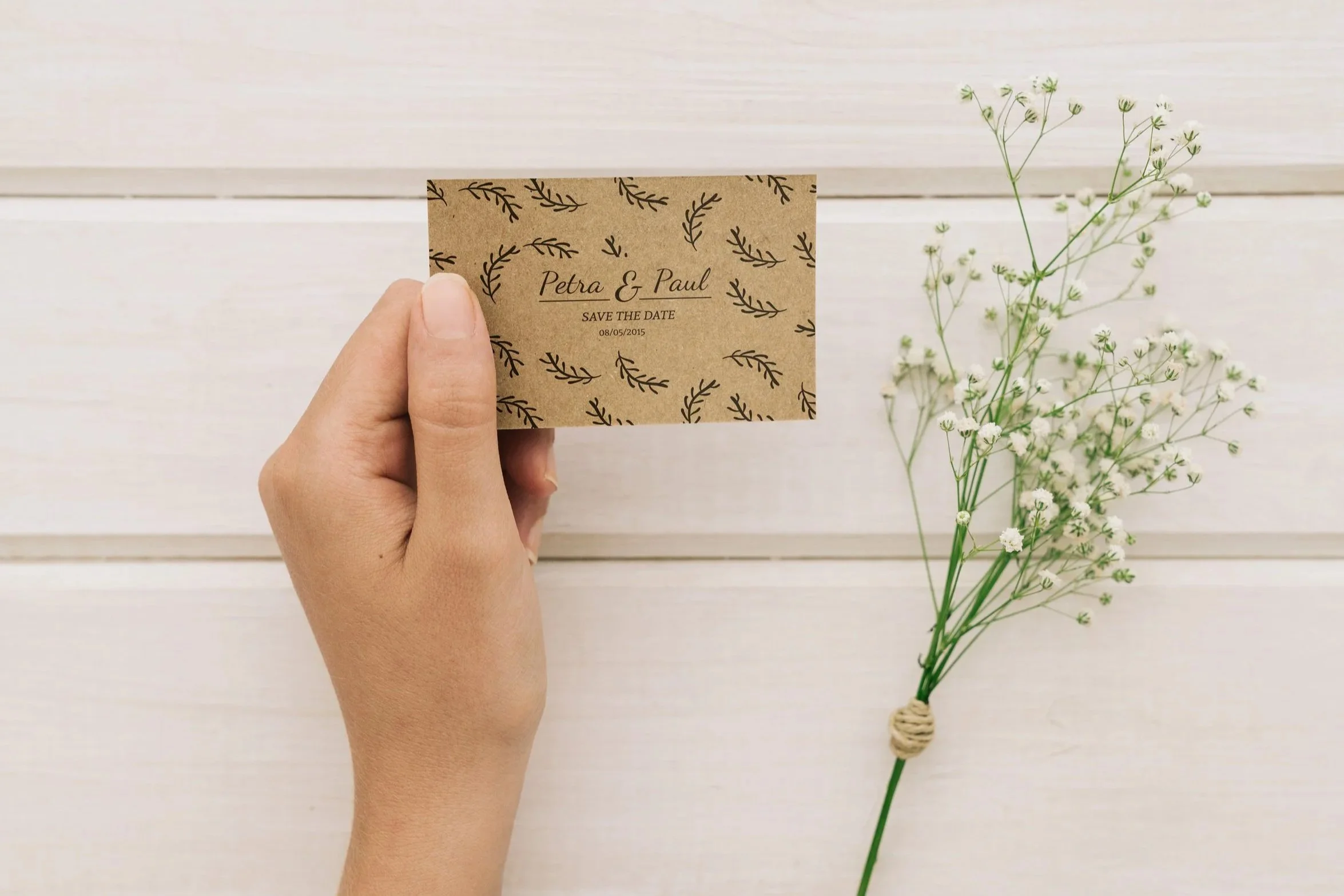 Hand holding a brown card with black leaf patterns and the text 'Petra & Paul, Save the Date, 08/05/2015' printed on it. A sprig of small white flowers is visible on the right side of the photo. The background is a white wooden surface.