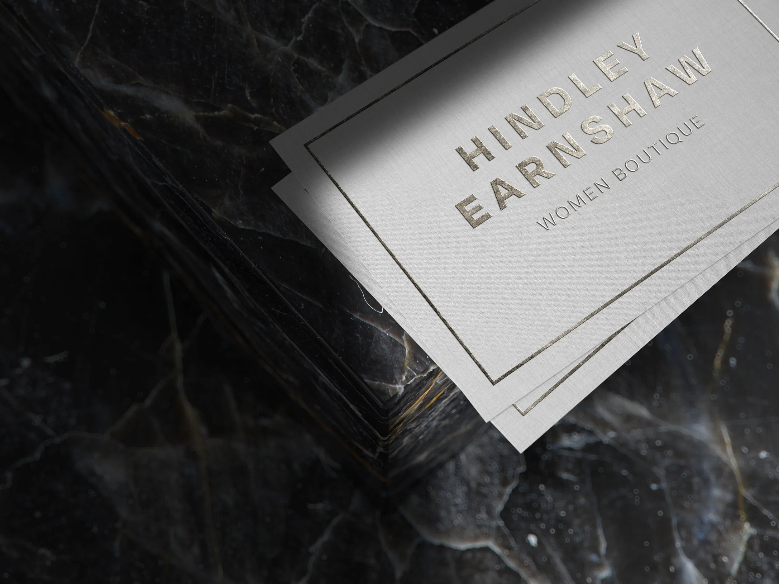 Close-up of a silver card with the text 'HINDLEY EARRINGS' and 'WOMEN BOUTIQUE' engraved on it, placed on a dark, marble-like surface.