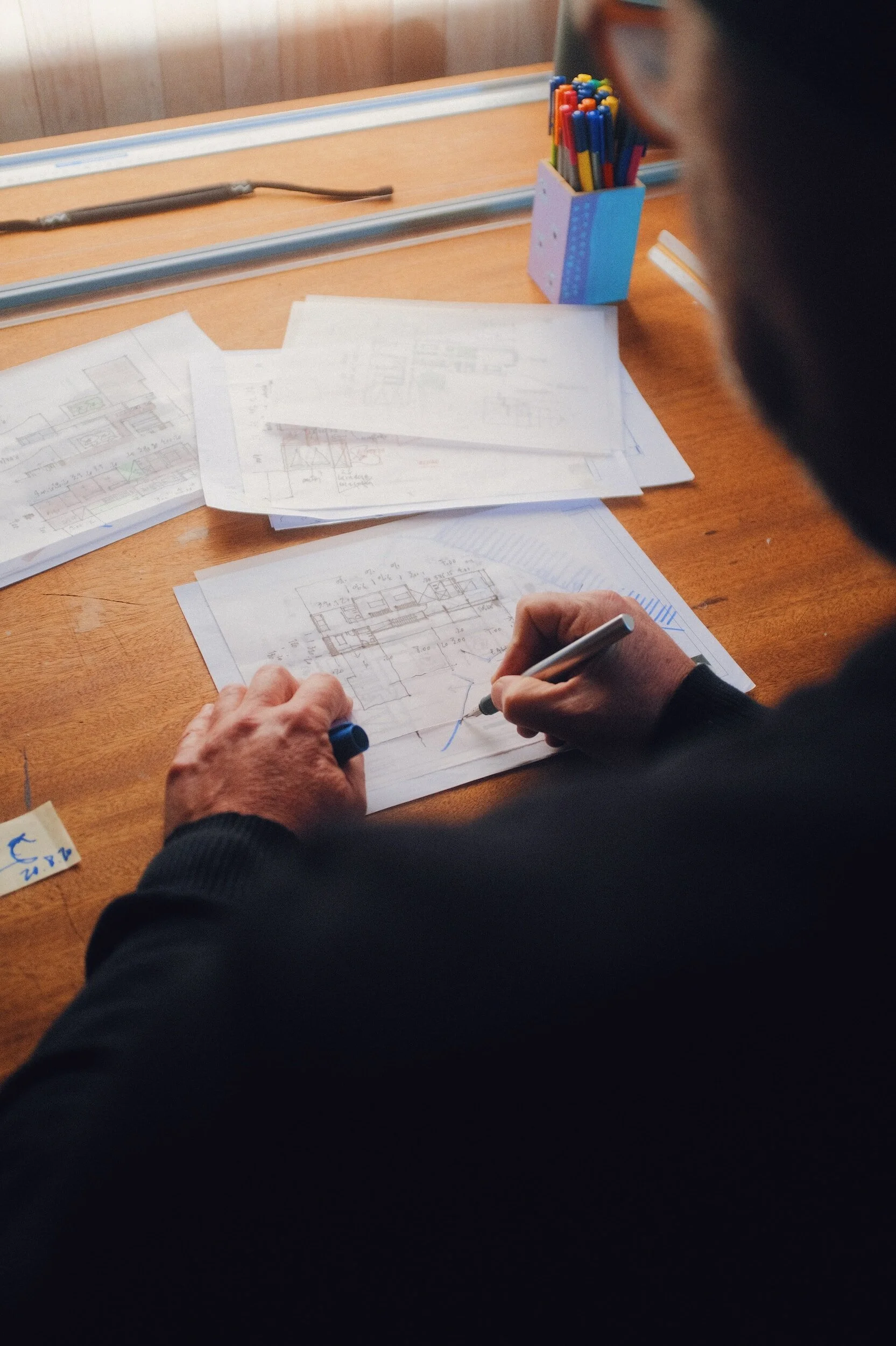 Person drawing architectural plans on paper at a wooden desk with colored pens in a holder.