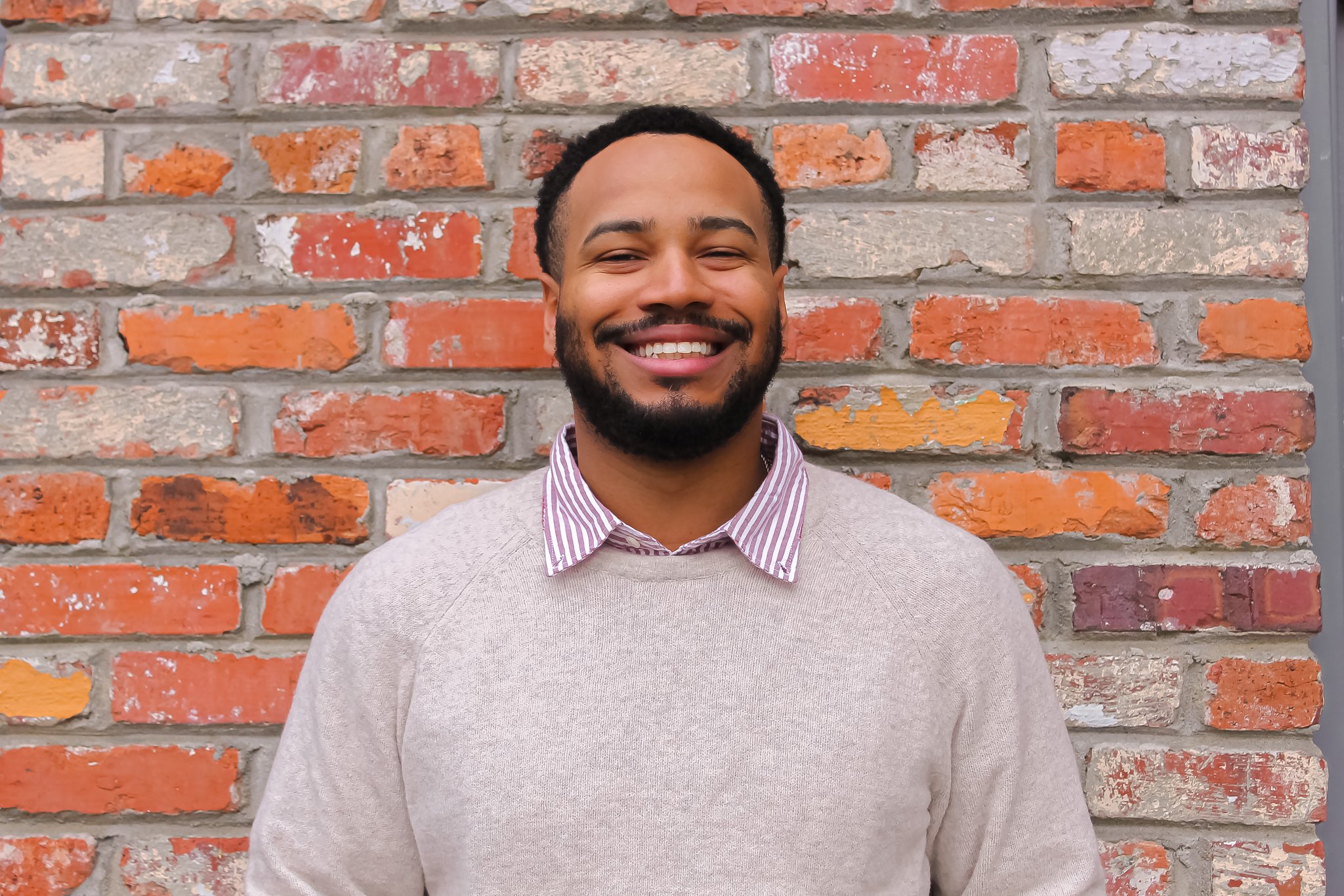 Person smiling in front of a brick wall wearing a light sweater and striped shirt.