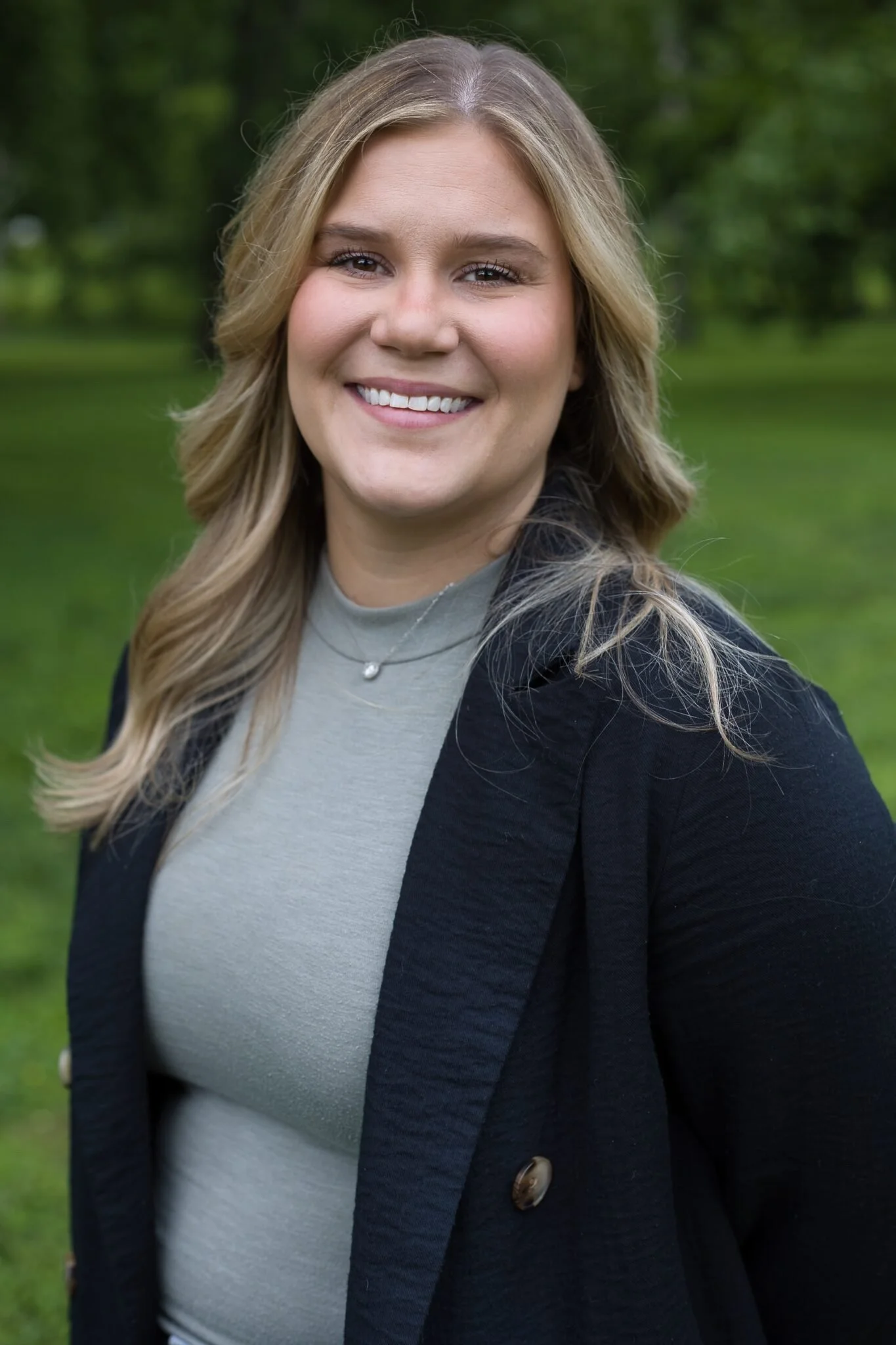 Woman smiling outdoors with green background, wearing a gray top and black coat.