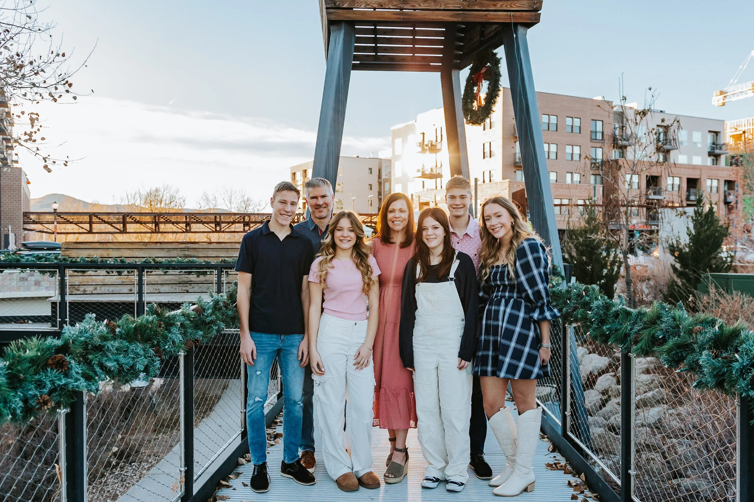 Group of seven people posing on a decorated bridge, with festive greenery and outdoor buildings in the background, during daytime.