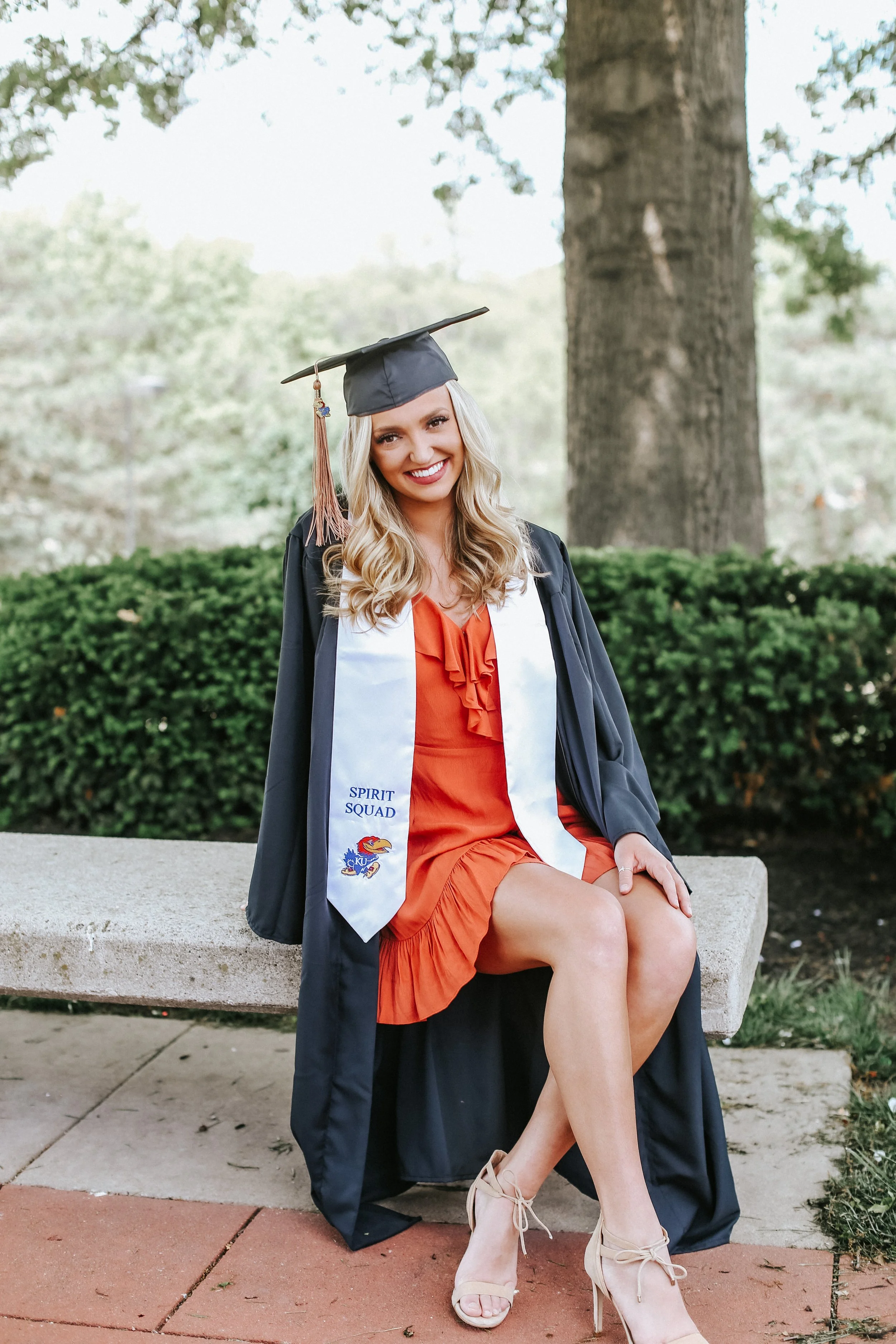 A young woman in a graduation cap and gown sits on a bench outdoors, wearing a red dress and a "Spirit Squad" sash.