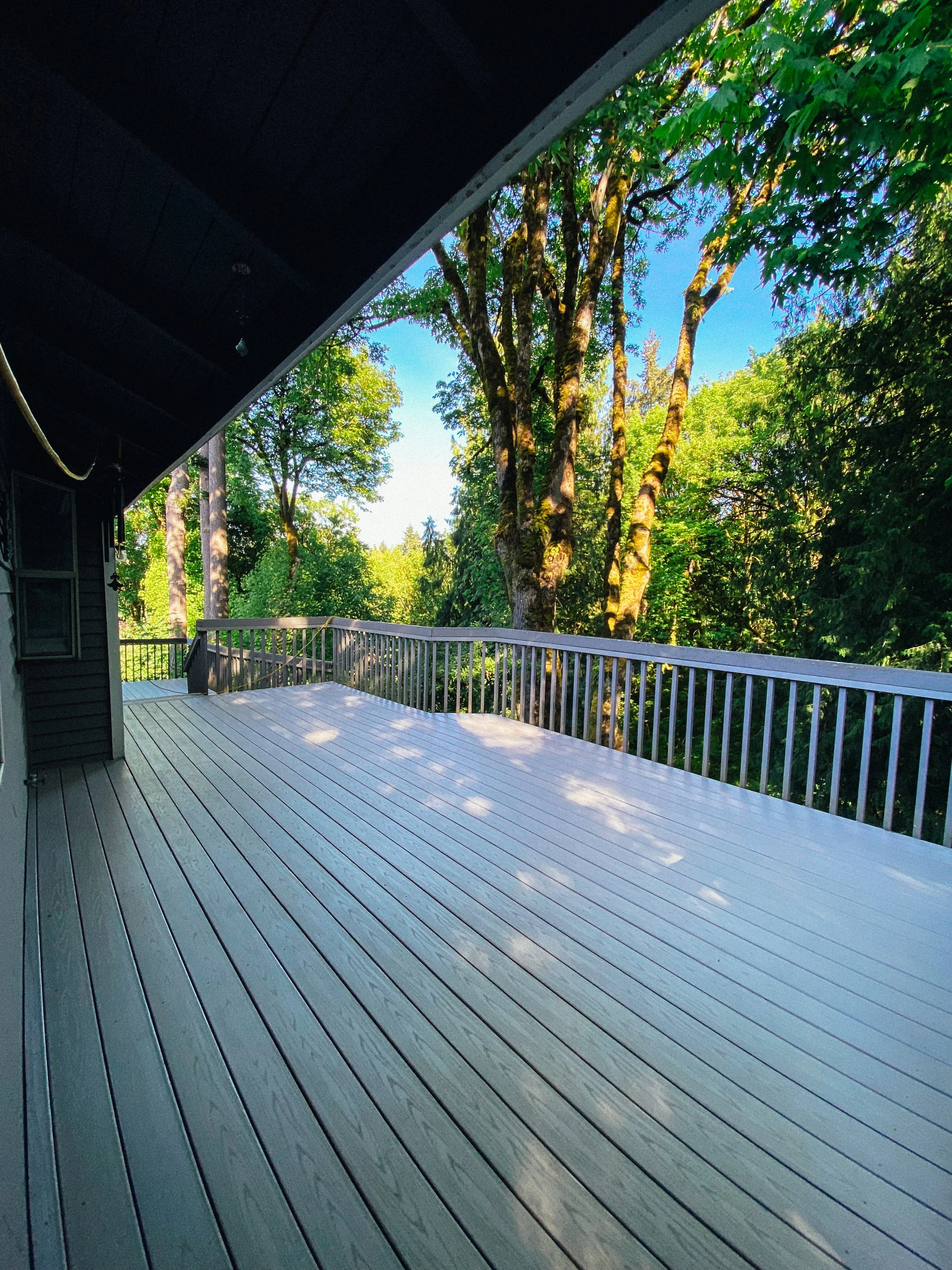 Wooden deck with white railing overlooking lush green trees under a clear blue sky.