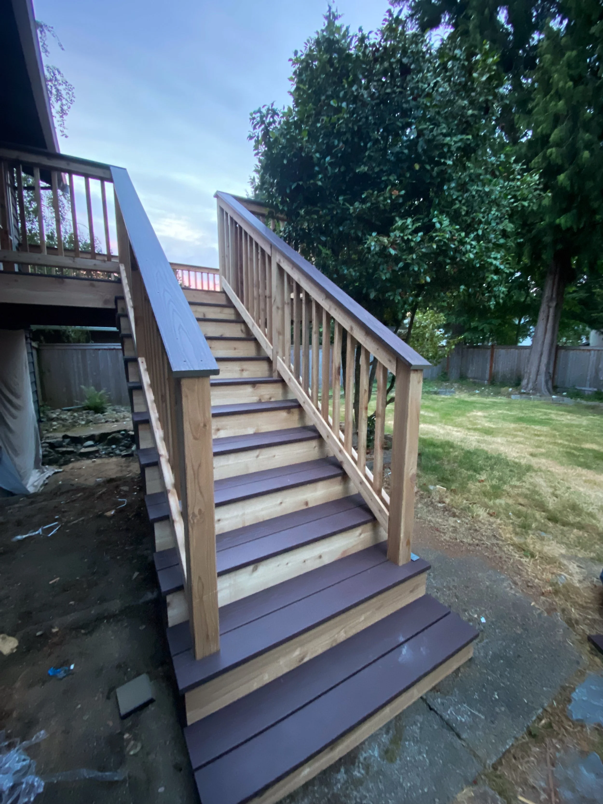 Newly built outdoor wooden staircase with dark brown steps and matching handrails in a backyard, leading up to a deck with a railing, surrounded by trees and a grassy yard.