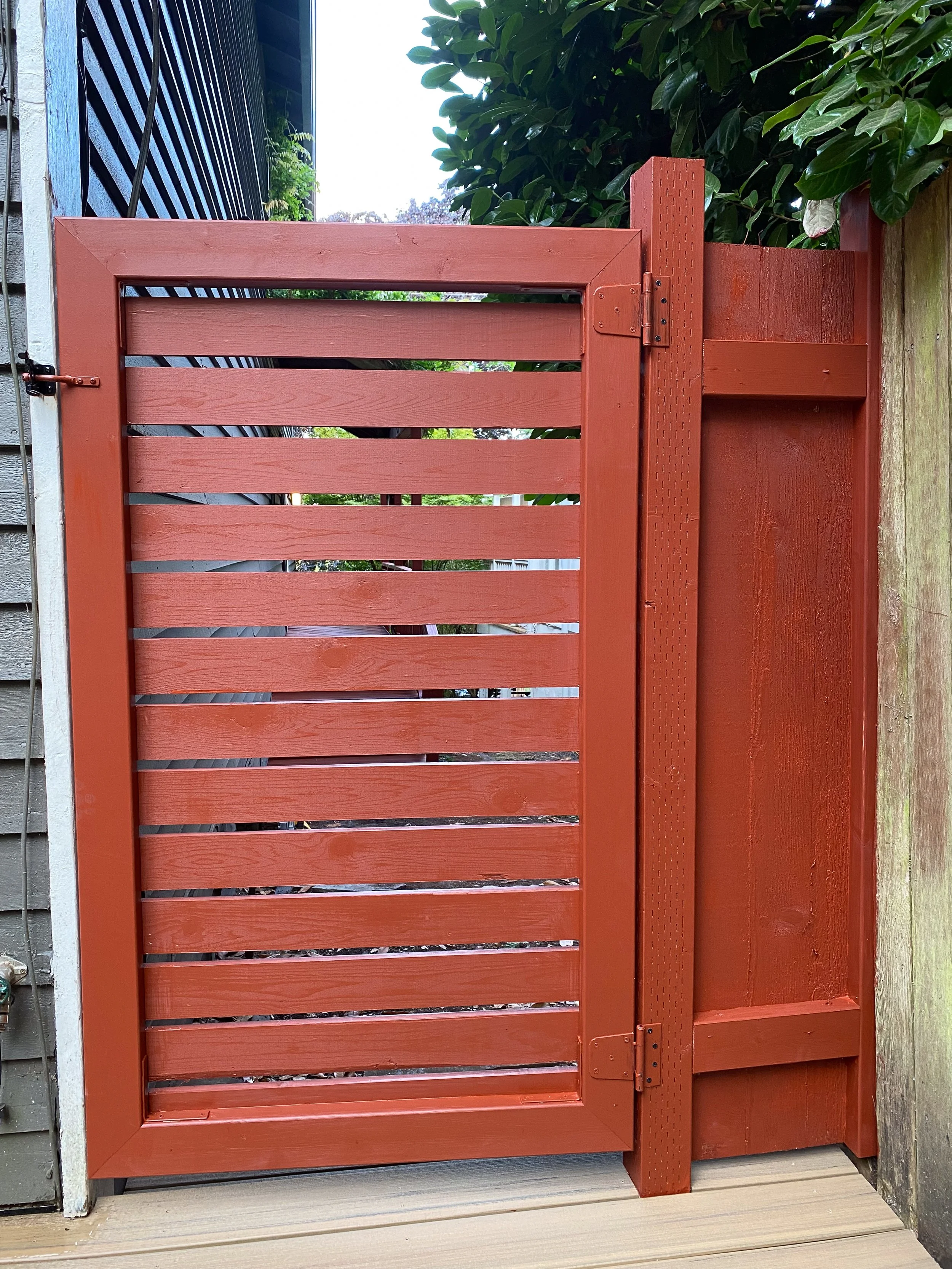 A red wooden gate with horizontal slats, mounted on a post, next to a wooden deck and a wall of a house, with a large leafy green shrub next to it.