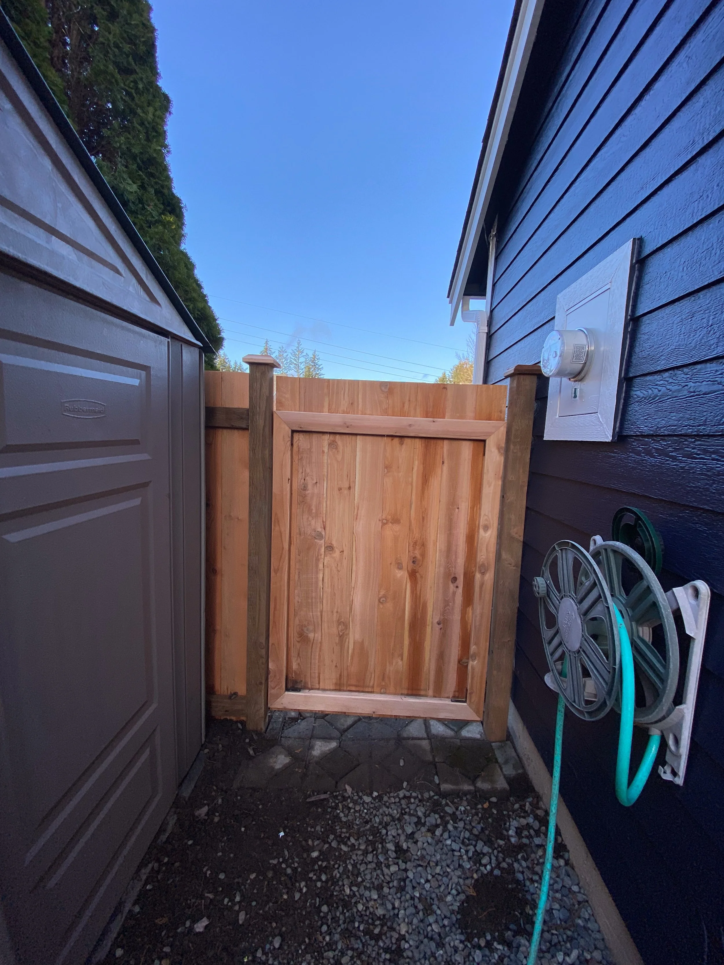 A newly installed wooden privacy gate between a gray storage shed and the dark blue house exterior with a garden hose reel attached to the wall, located in a small outdoor space with a clear blue sky.