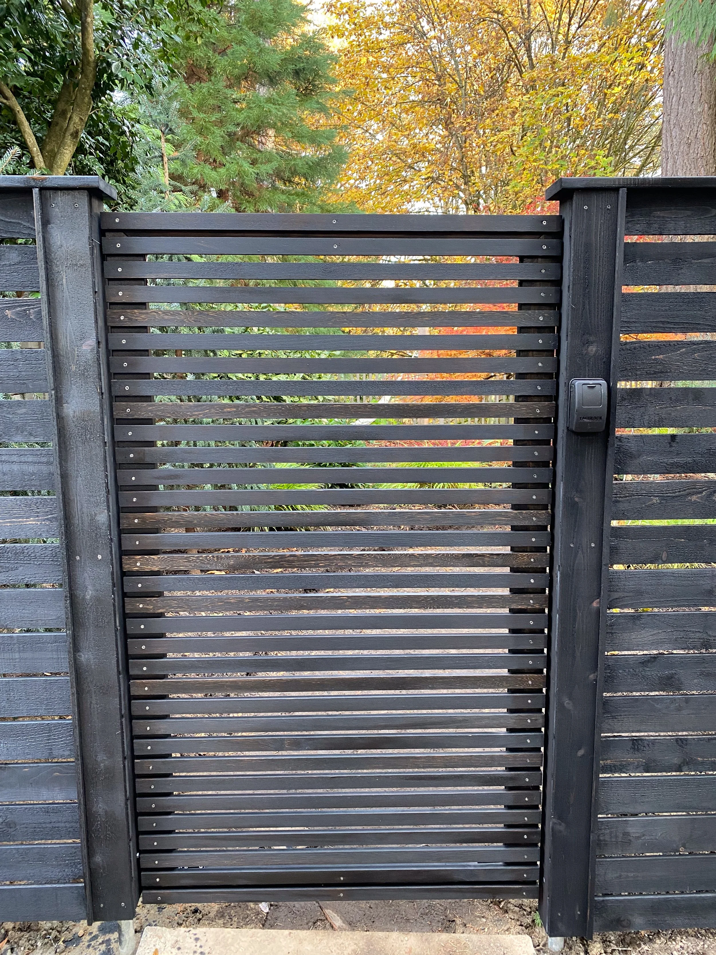 A black wooden slat fence with horizontal slats, equipped with an electronic gate opener, set outdoors with trees showing fall foliage in the background.