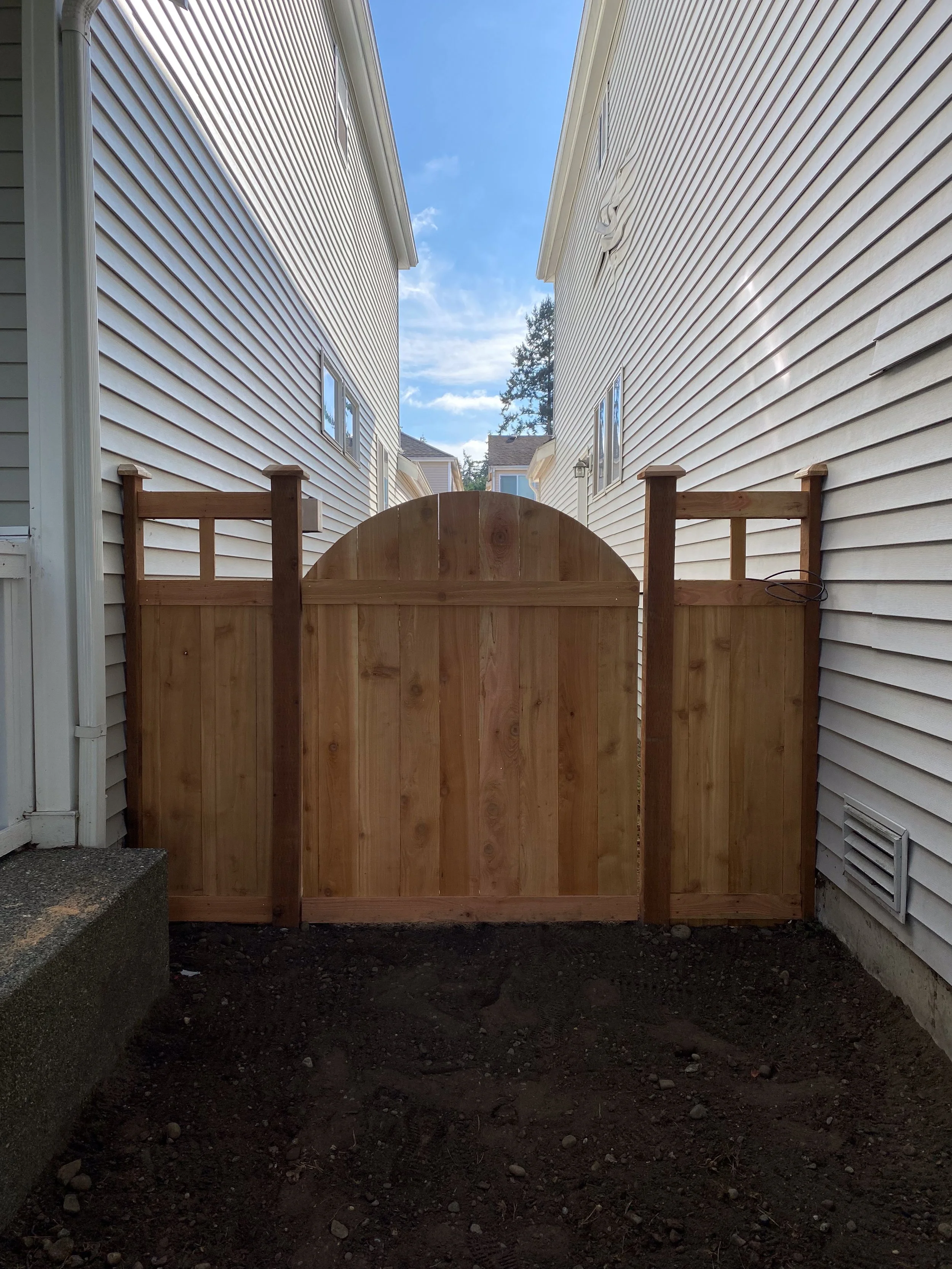 A wooden gate between two white houses with vinyl siding, a clear blue sky, and trees in the background.