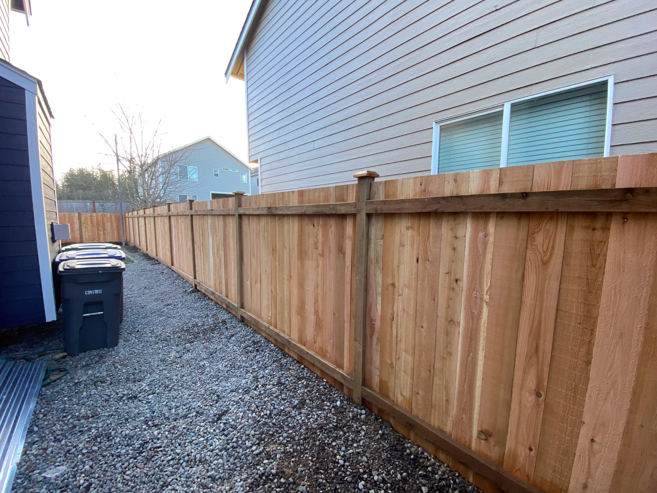 A backyard with a newly installed wooden fence, gravel ground, and neighboring houses visible in the background.