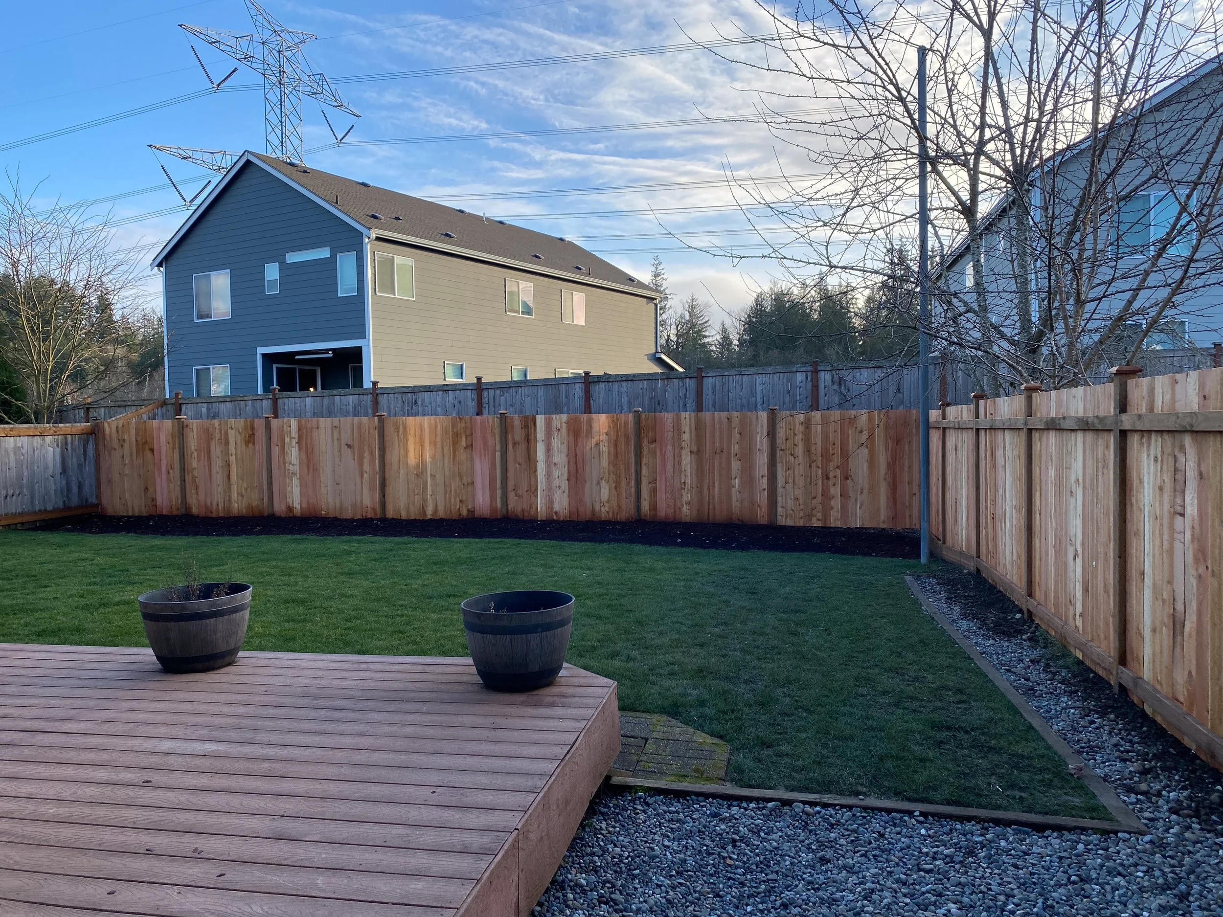 A backyard with a wooden deck, two large planters, a lawn, a new wooden fence, and houses in the background under a blue sky with clouds.