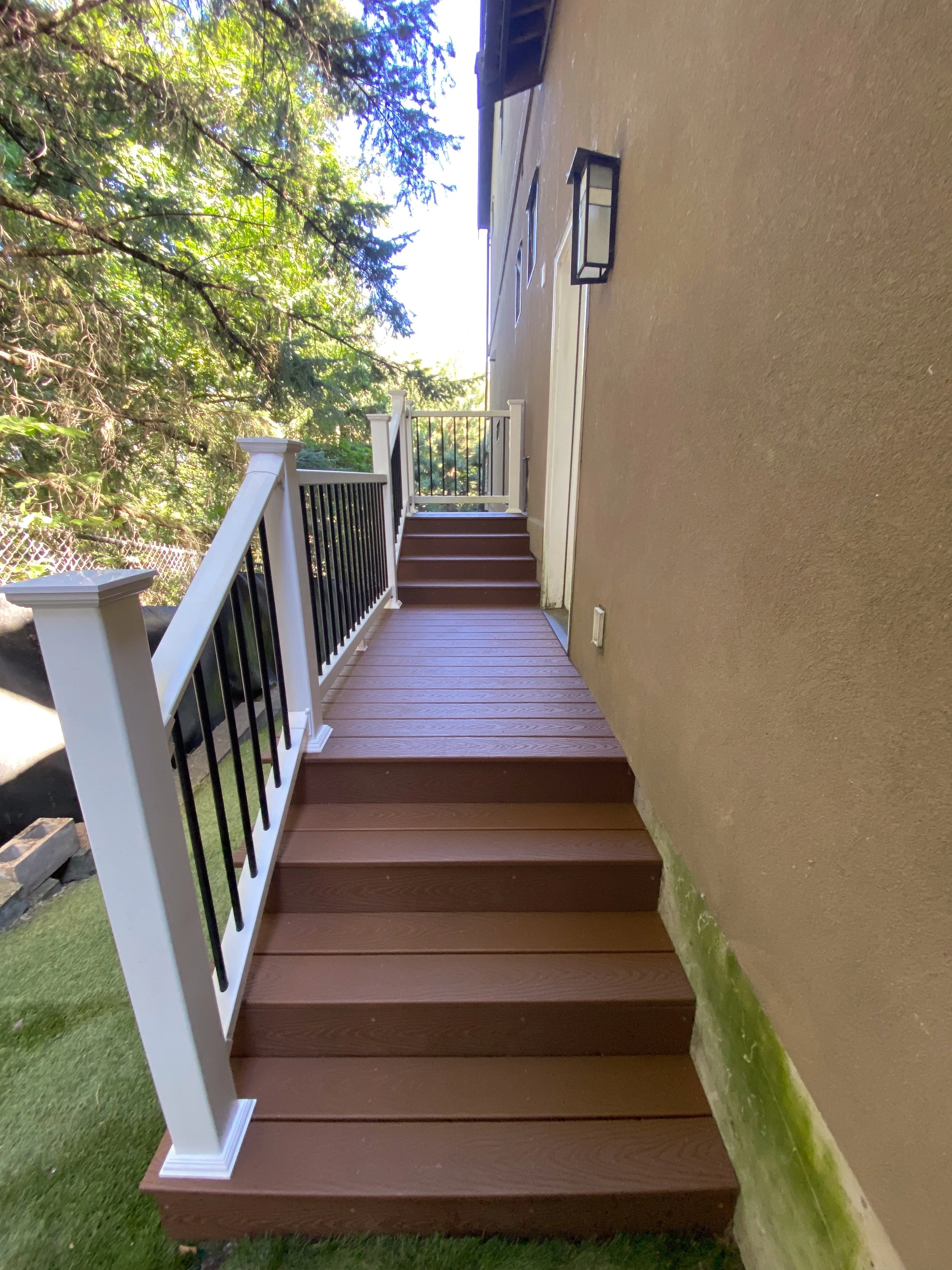 Outdoor wooden deck attached to beige house, with steps leading up to a door, black railing, and surrounded by trees.