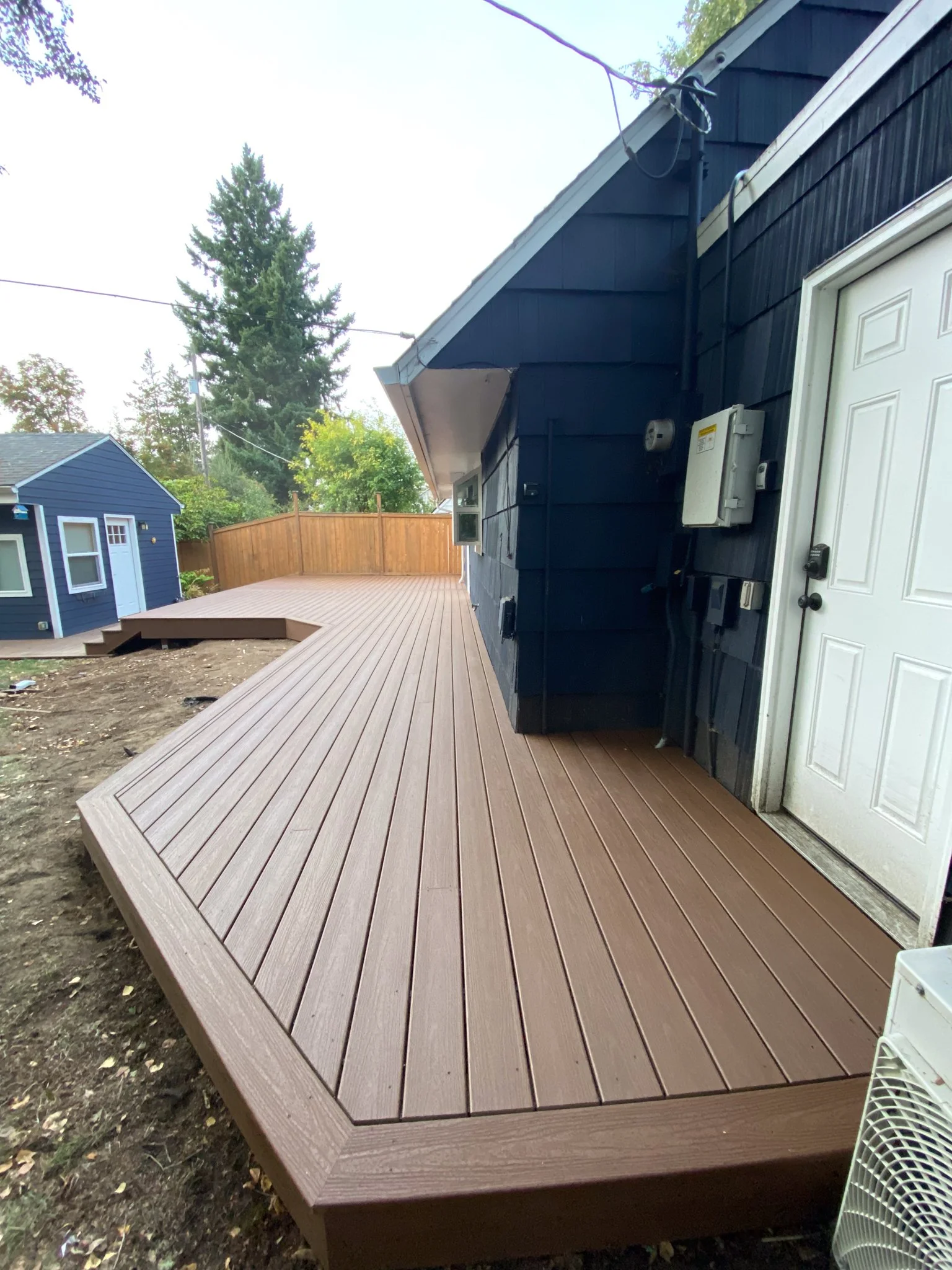 Newly built wooden deck attached to a house with a dark blue exterior and a white door, with a neighboring blue house in the background.