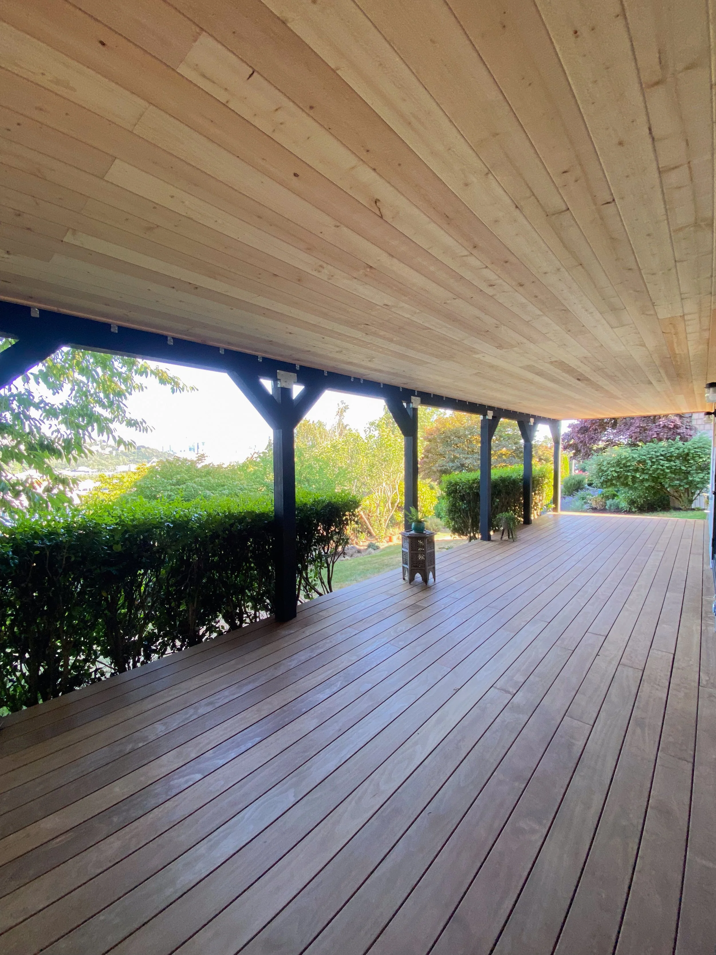 Spacious wooden porch with a ceiling, supported by black beams, overlooking a lush garden with bushes and trees.