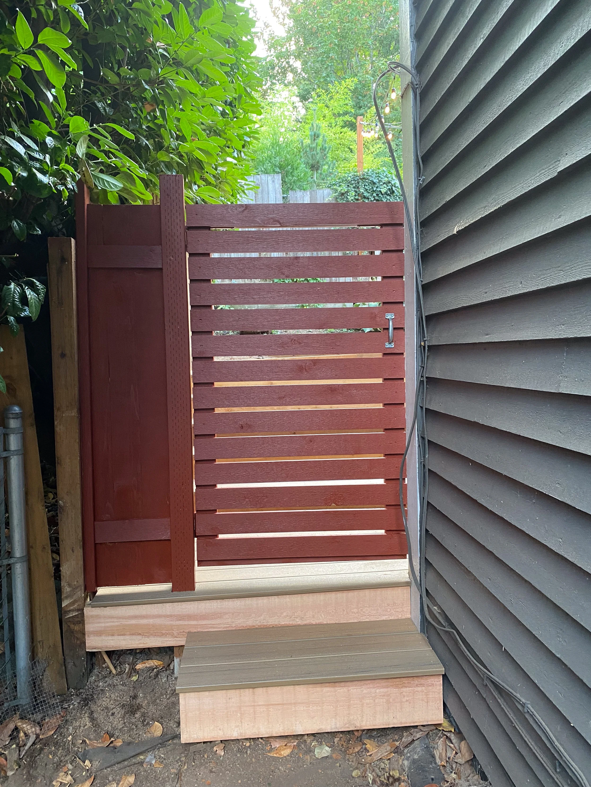 A small wooden deck with a reddish-brown privacy fence, attached to a house with gray horizontal siding, surrounded by green trees and bushes.