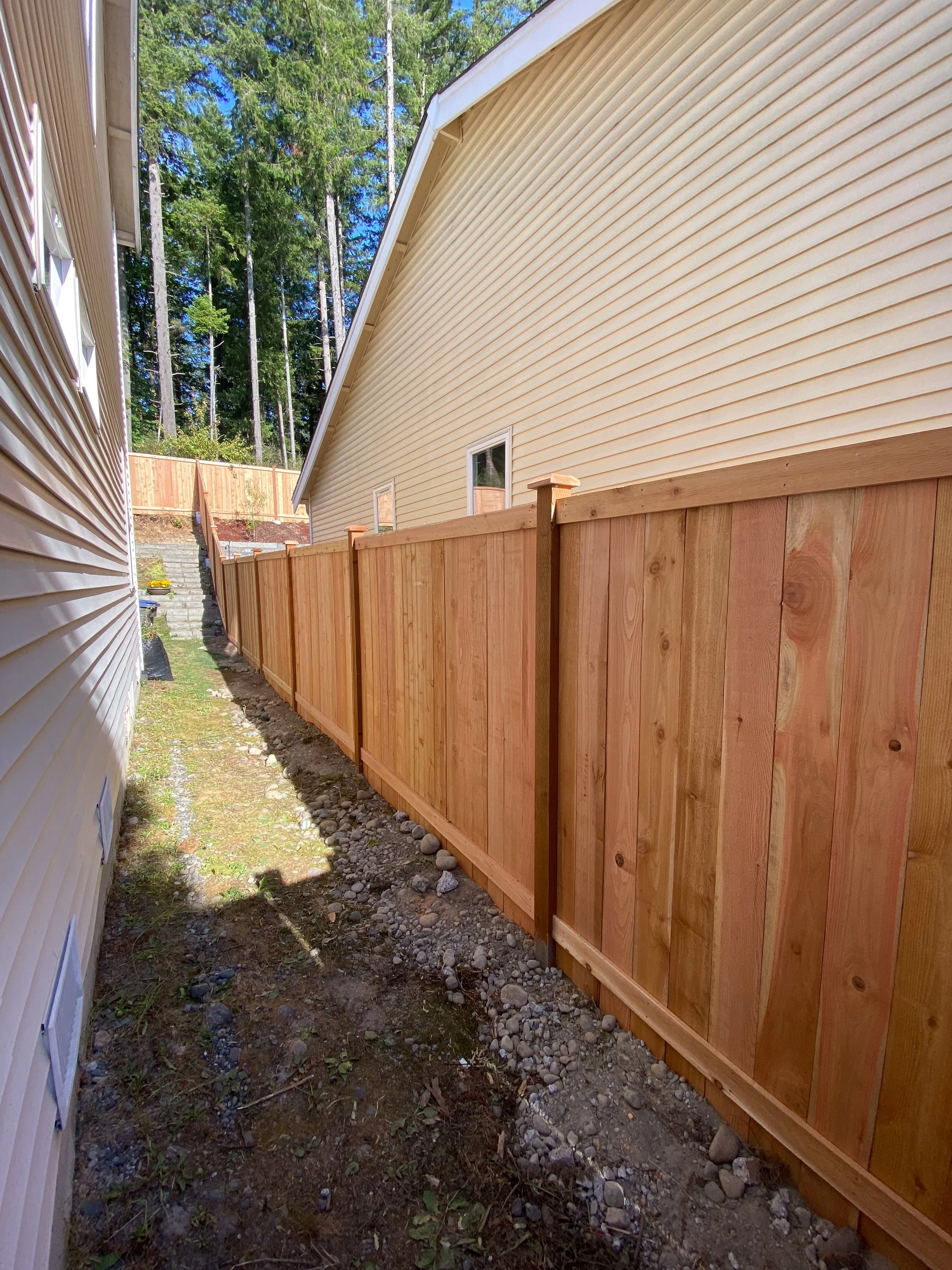 View of a newly built wooden fence running alongside a house with beige vinyl siding, with a gravel and dirt pathway in between, and trees visible in the background.