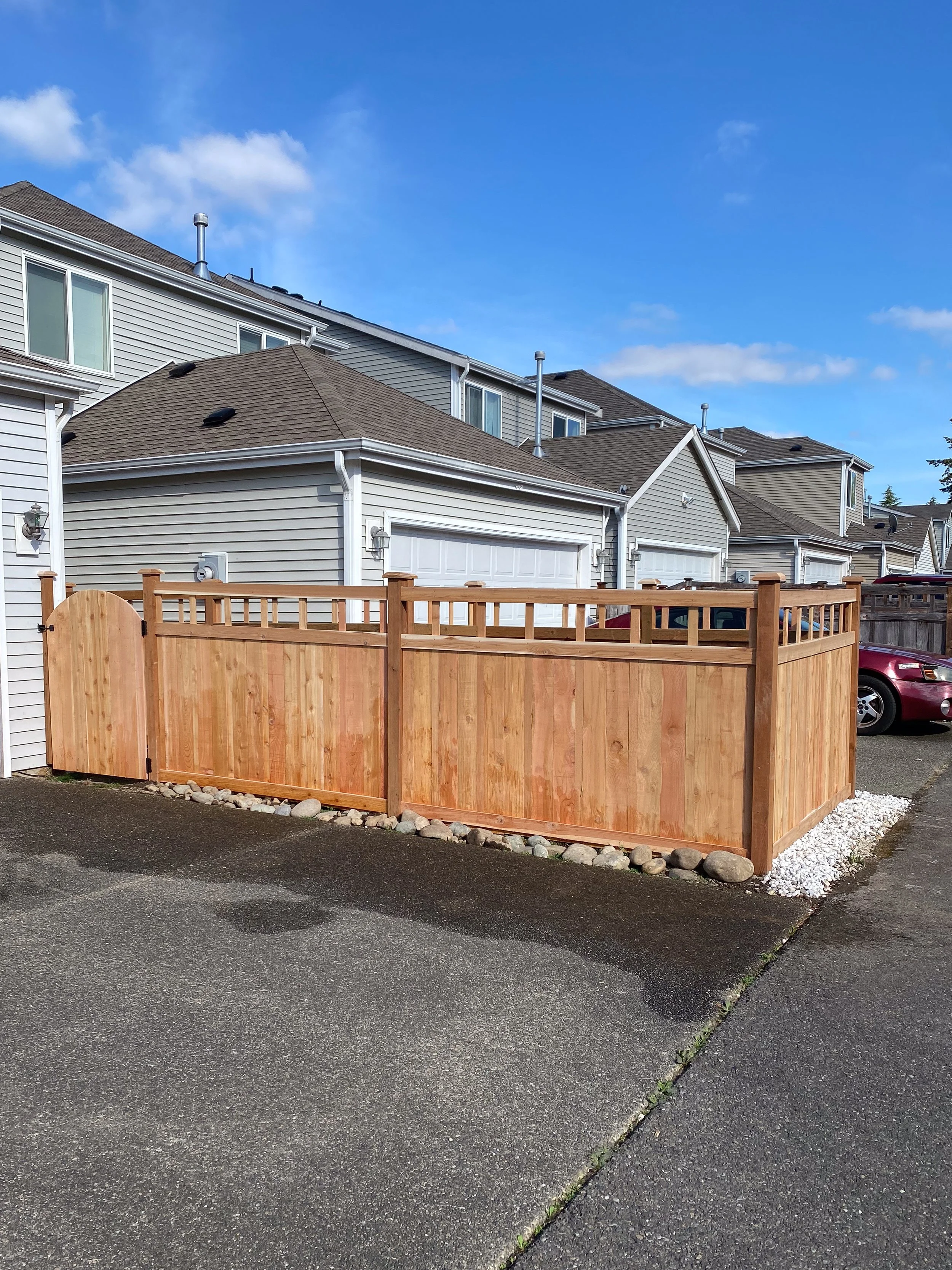 A newly built wooden fence with decorative top railing surrounds a front yard in a residential neighborhood. There are townhouses in the background with gray siding and white trim, and a clear blue sky overhead.