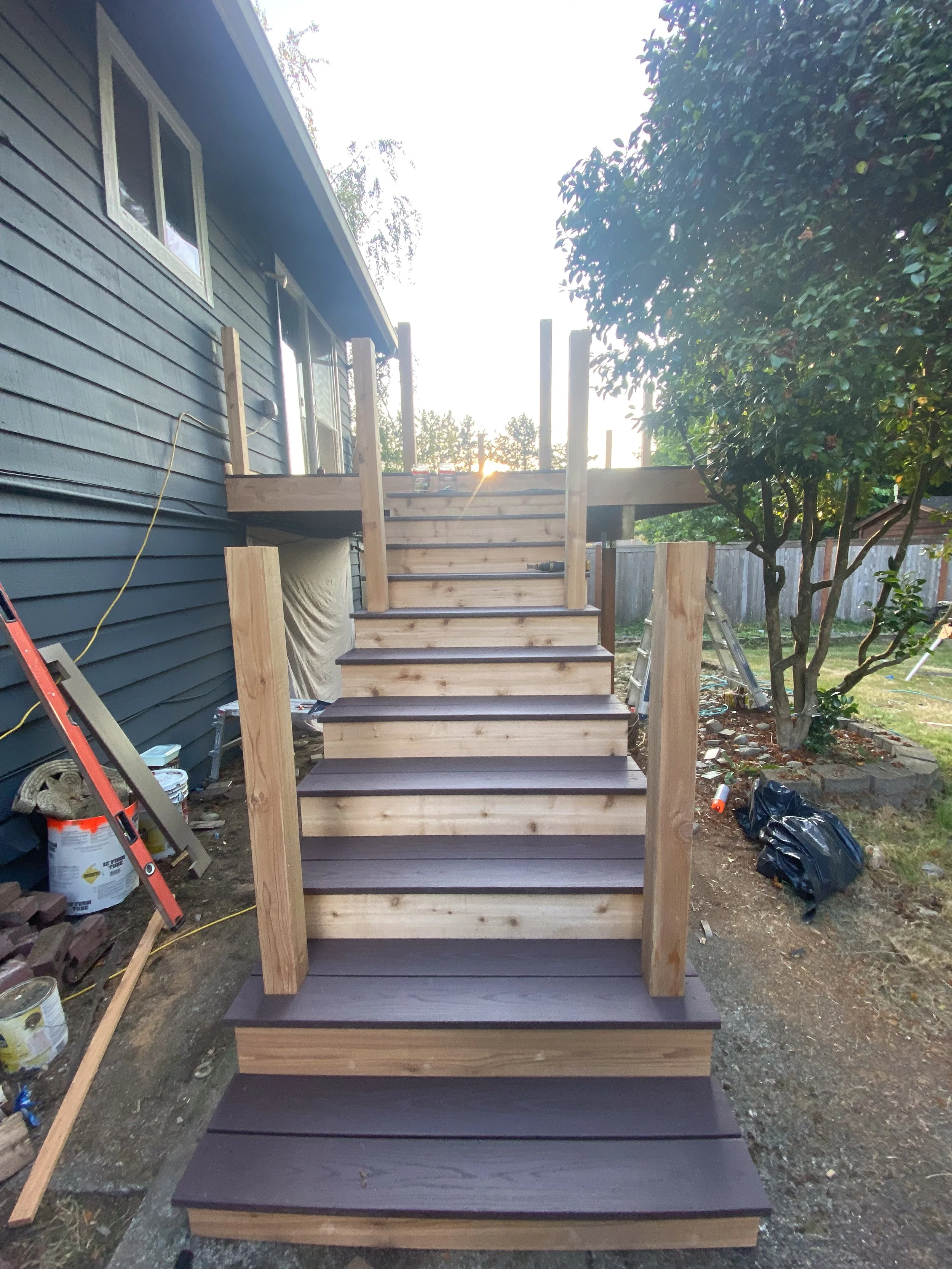 Wooden staircase under construction outside a house with black siding, a tree on the right, and construction tools and materials around.