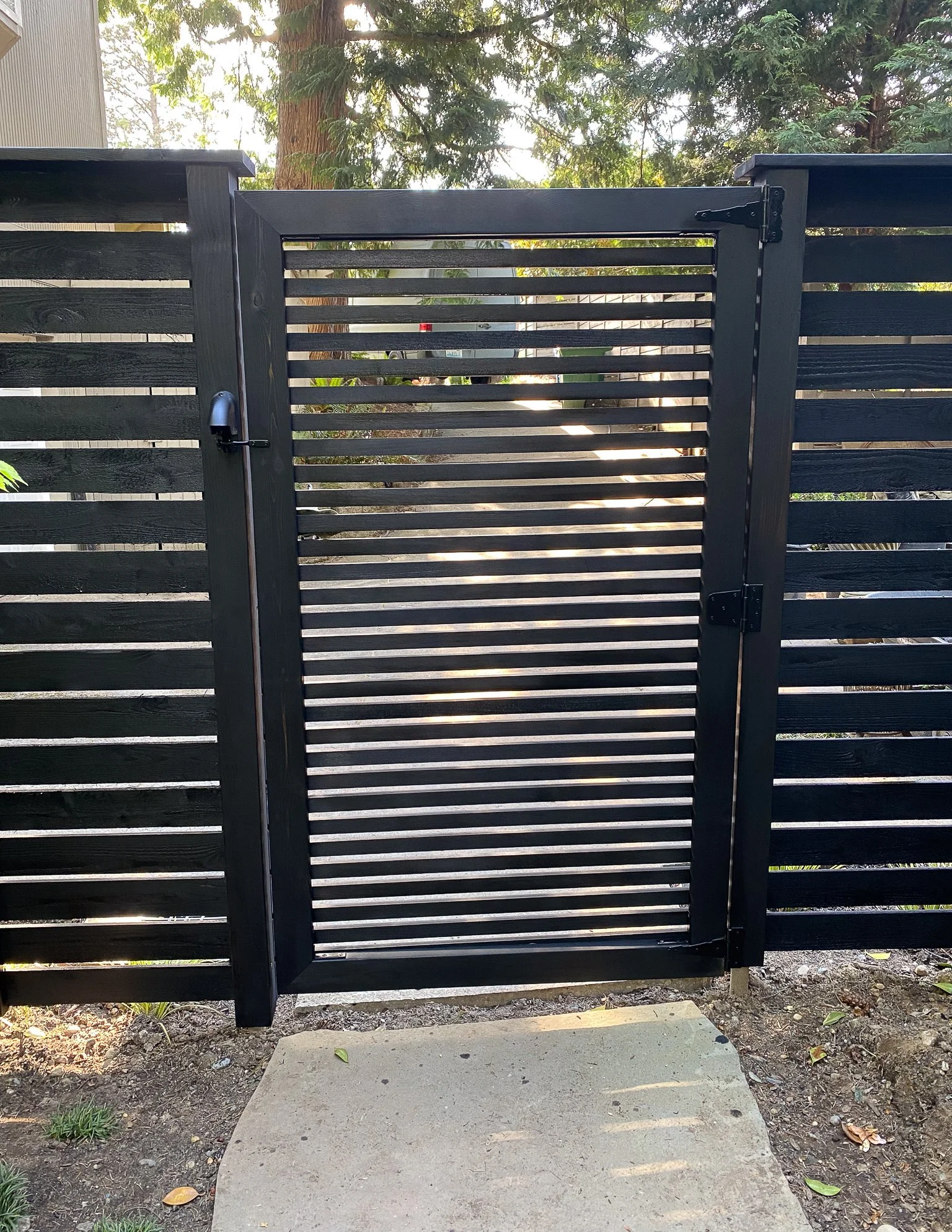 Black louvered gate with a latch, part of a black wooden fence, and trees visible behind it.