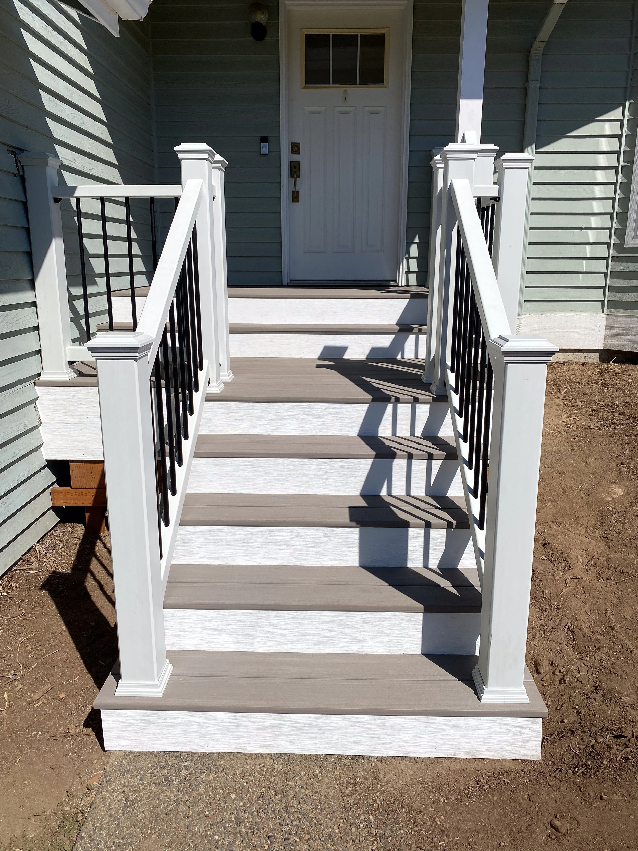 Newly built front porch stairs with white and gray color, black metal railings, leading to a white door with a window pane above, attached to a grey house with horizontal siding.
