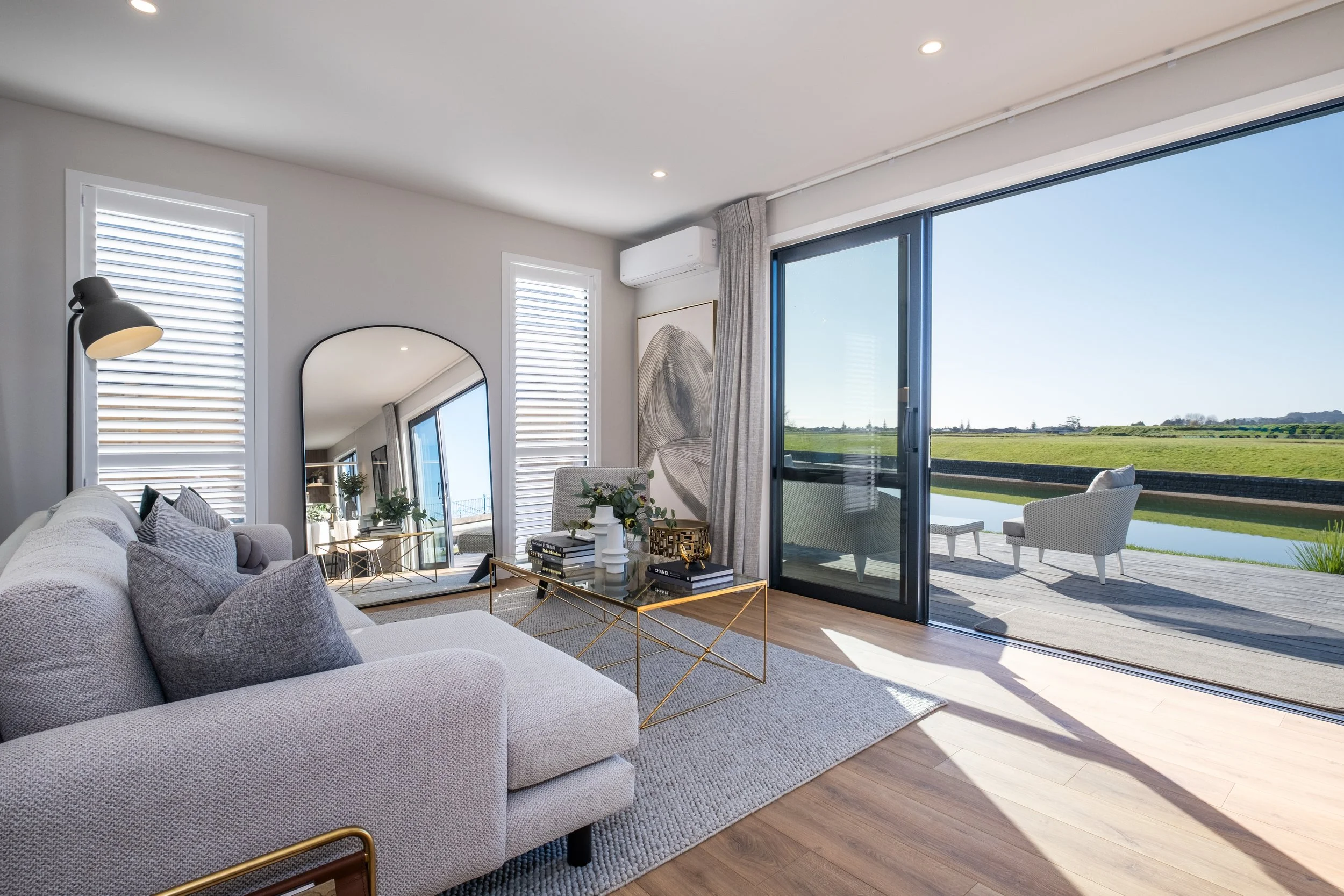 Living room with beige sofa, gray pillows, glass coffee table, large mirror, and sliding glass door leading to balcony with outdoor seating and river view