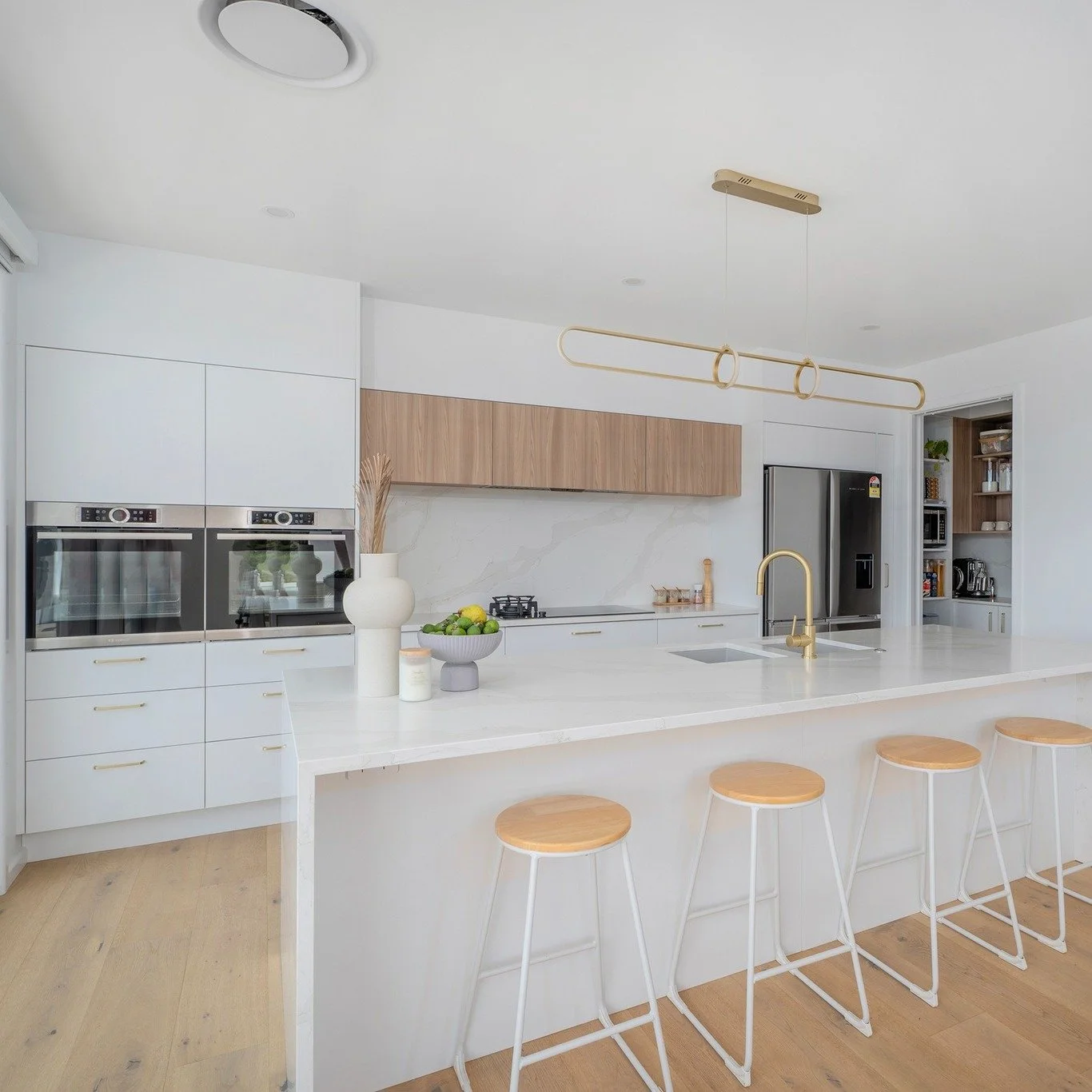 Modern kitchen with a white island, four wooden barstools, built-in ovens, a stainless steel refrigerator, and open shelving with kitchenware.
