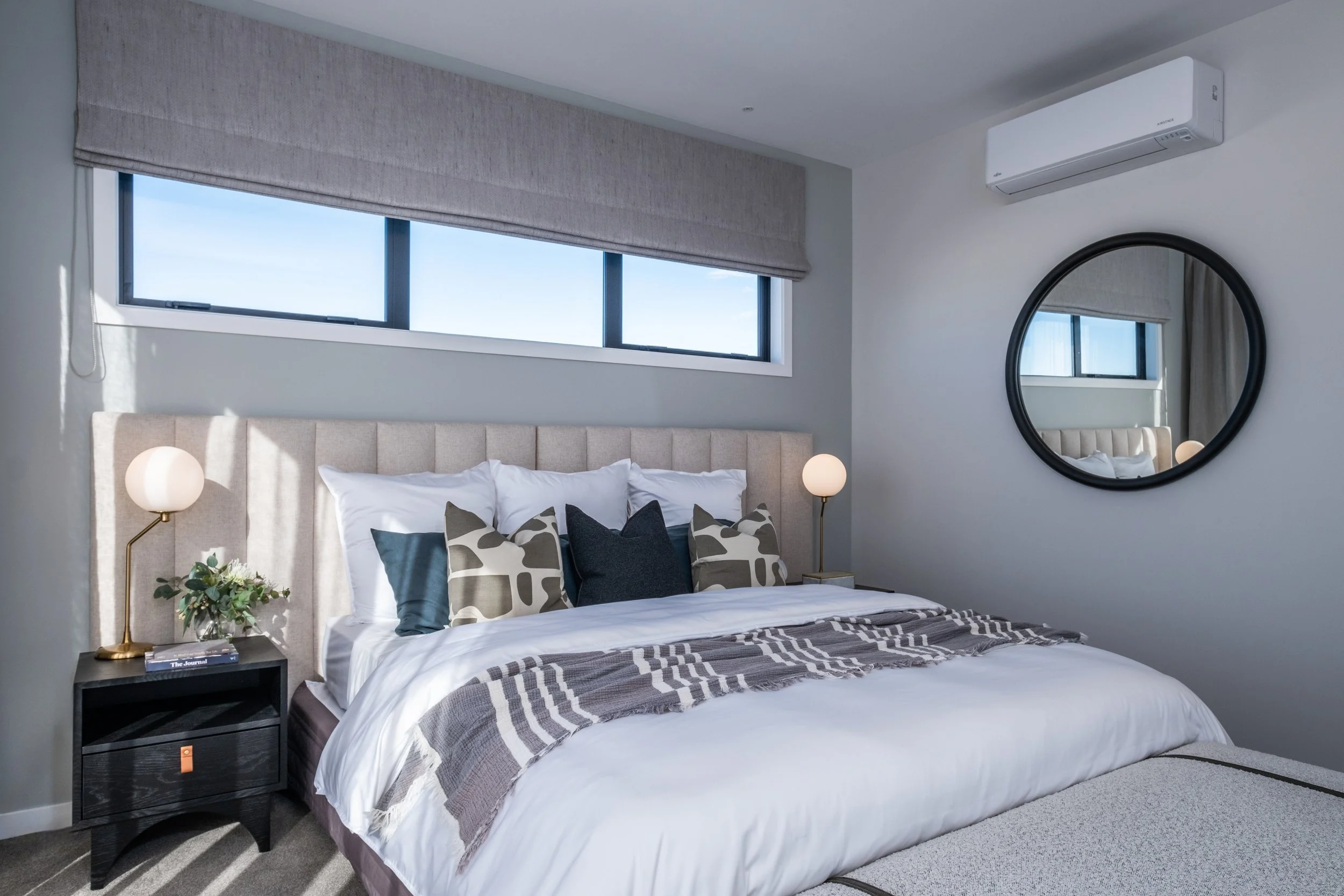 A modern bedroom with a large bed, decorative pillows, bedside lamps, a window with a gray Roman shade, a round mirror on the wall, and a split air conditioning unit.