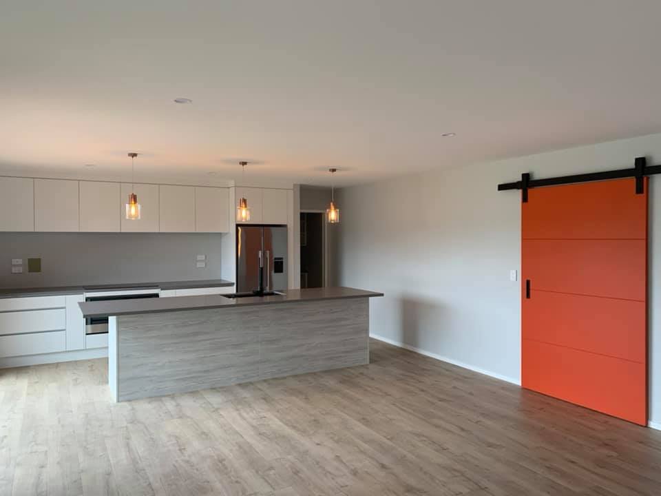 Empty modern kitchen with white cabinets, gray countertops, a wooden island, stainless steel refrigerator, and a bright orange sliding barn door.