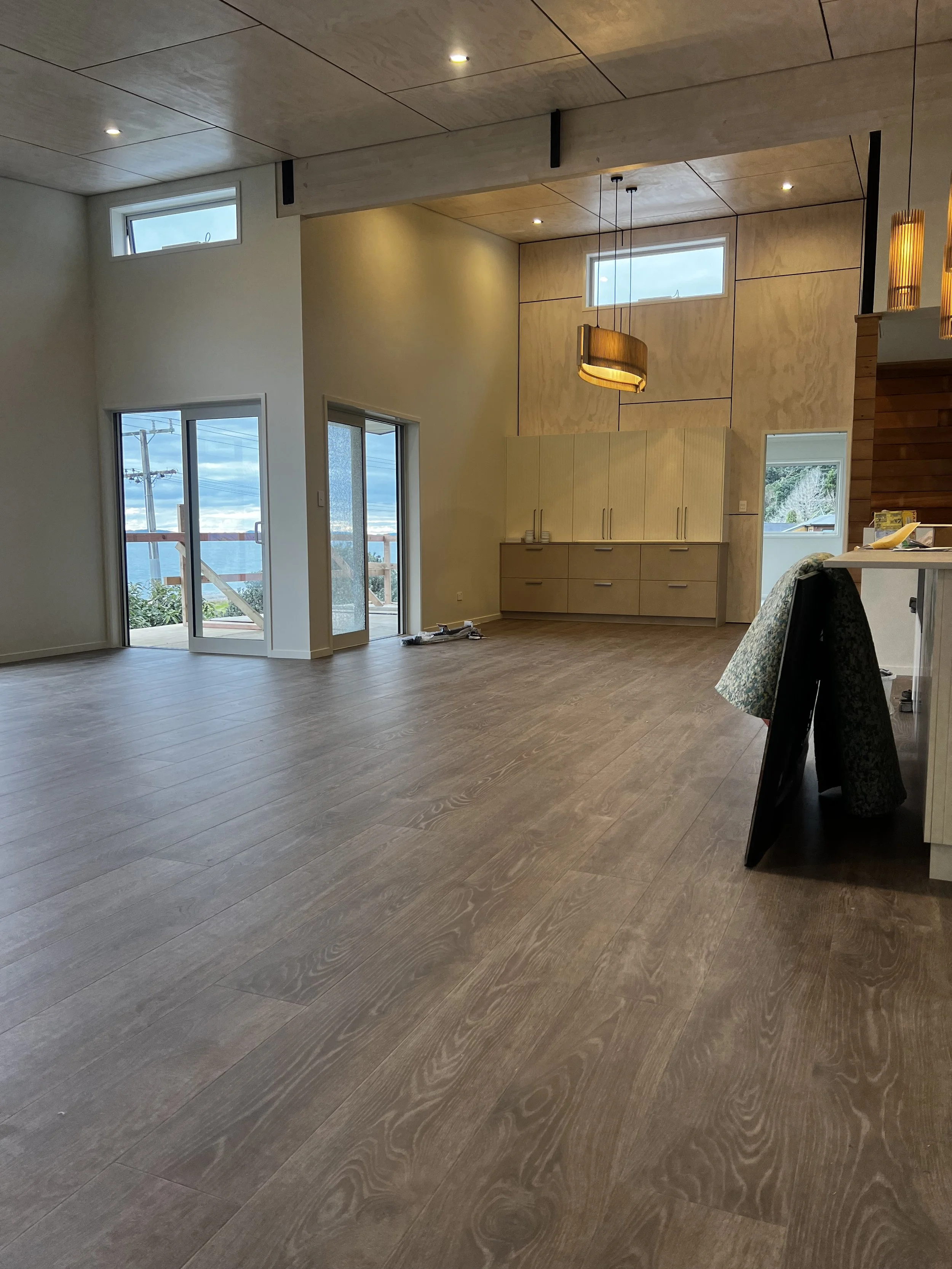 Empty modern living room with wooden floors, large windows, and a sliding door leading outside, with built-in cabinets and hanging lights.