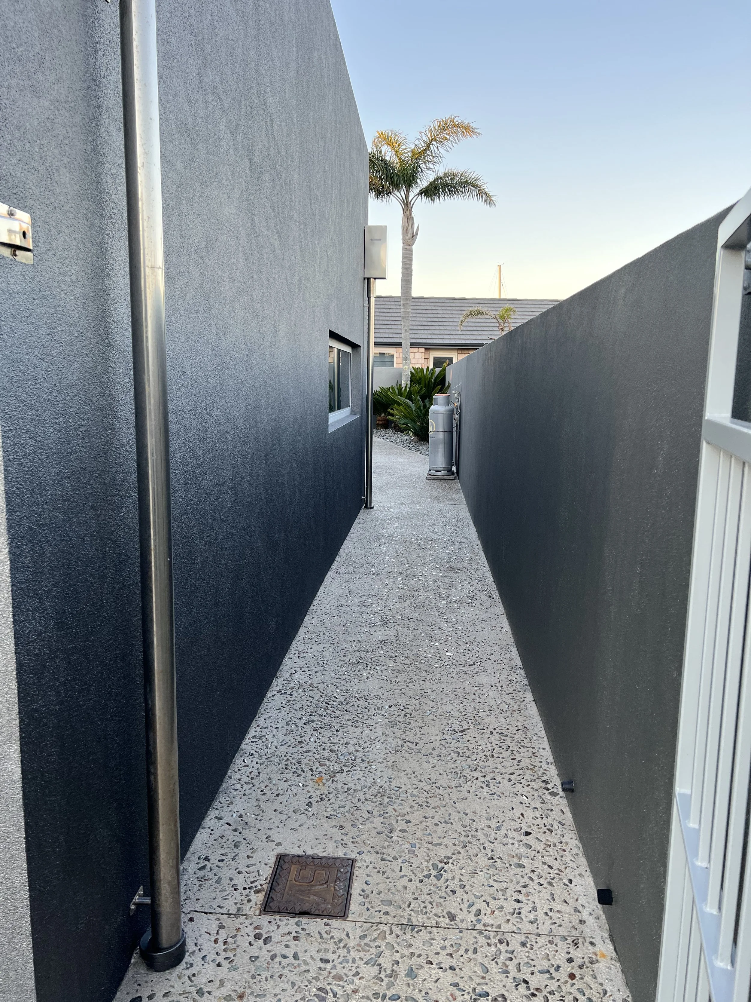 A narrow outdoor alleyway between two modern dark-colored walls, with a palm tree and a small garden in the background, and a trash can near the end of the alley.