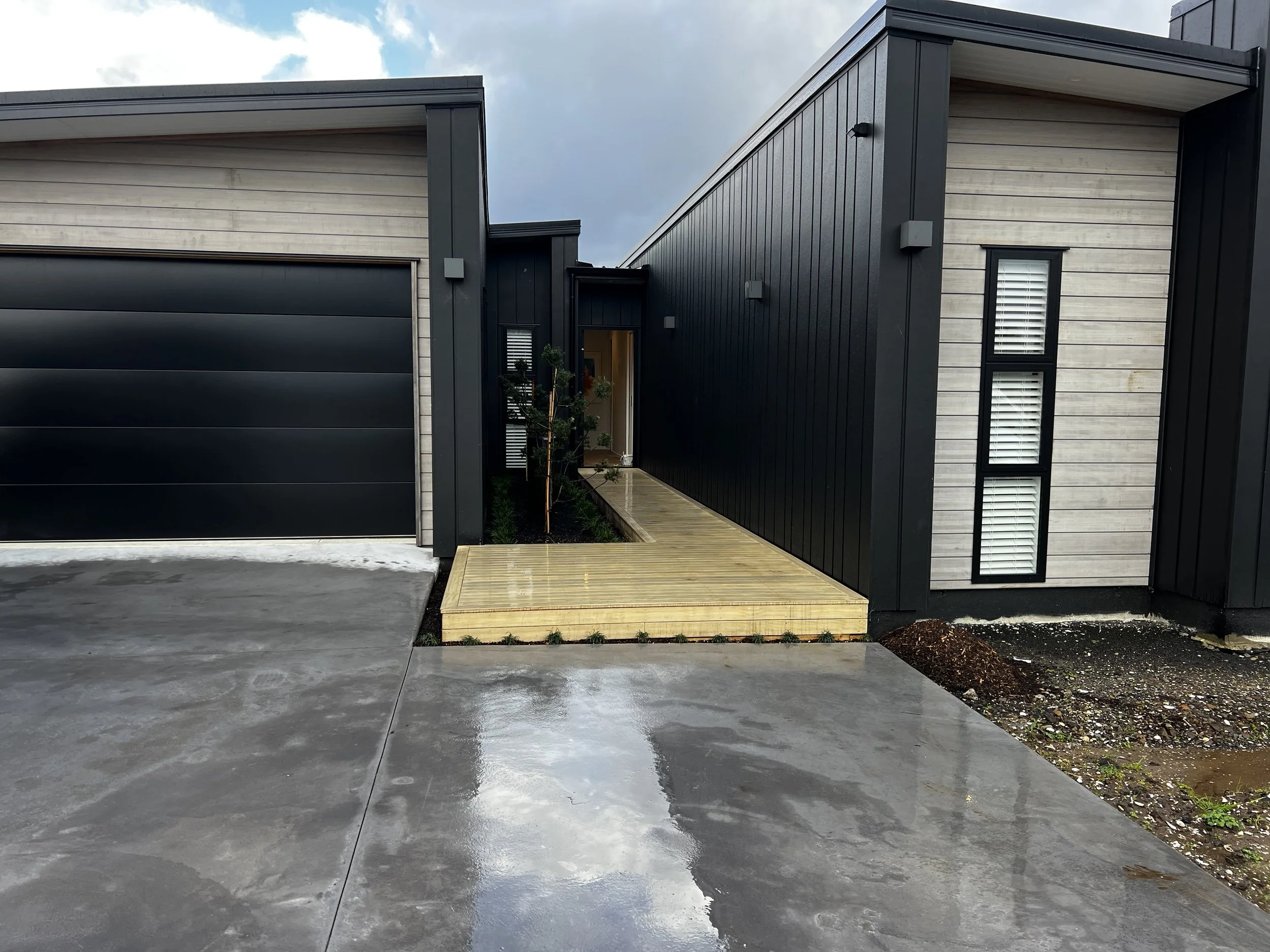 Modern house exterior with black and light wood siding, a dark garage door, a narrow wooden pathway, and a small tree near the entrance.