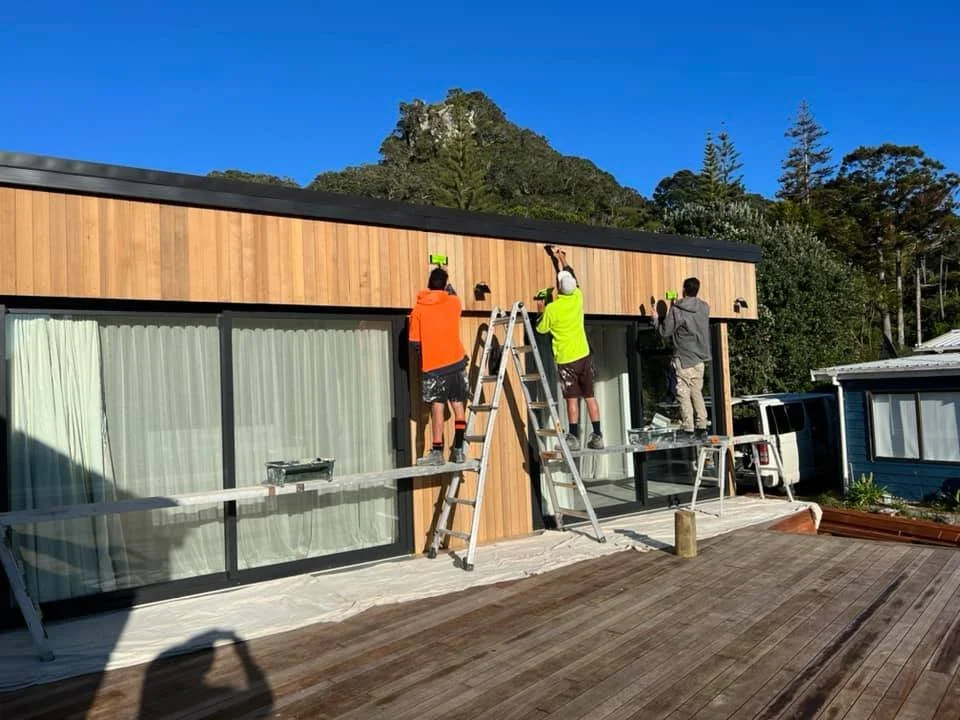 Three people working on installing lighting fixtures on the exterior of a modern house with large glass doors and wooden paneling, with a mountain and trees in the background.